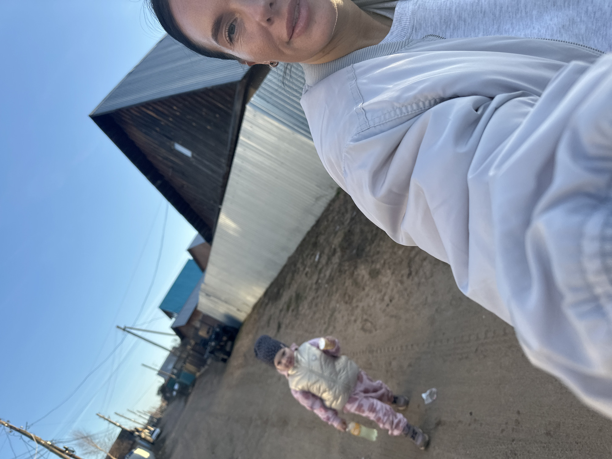 This outdoor selfie-style image is set in what appears to be a rural or semi-rural village, specifically identified as Olkhovka, Russia. The photo is taken from a high angle, looking downwards and across an unpaved road or dirt path.
In the immediate foreground, to the right, is an adult female who is taking the picture. Only her upper face (right eye, nose, mouth) and part of her torso are visible. She has dark hair and a slight, pleasant expression. She is wearing a light grey jacket over a lighter grey sweatshirt.
Further down the dirt path, on the left side of the frame, stands a child. The child is wearing a dark knit hat, a light-colored puffer vest, and patterned pink or purple pants. They are holding a clear plastic bottle in one hand. The child's face is slightly blurry but seems to be looking towards the camera.
The background shows a row of modest buildings lining the unpaved road, which stretches into the distance. Some buildings feature dark wooden siding, while others are constructed with corrugated metal. One building has a distinct blue roof. Multiple utility poles with strung wires are visible alongside the path. The sky is clear and bright blue, suggesting a sunny day, likely during late morning or early afternoon. The ground is dry and dusty, with visible tire tracks. No visible text is present in the image.