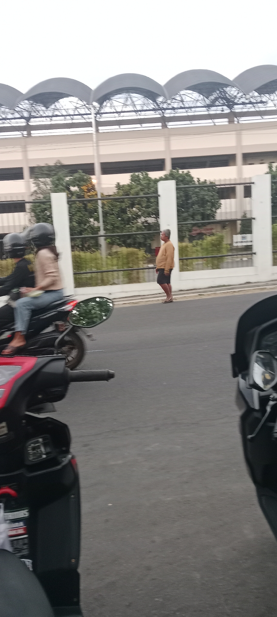 A medium-shot captures a street scene with several motorbikes and a lone pedestrian, likely in Ngemplak, Indonesia, under an overcast sky. In the foreground, the handlebars and front section of a red scooter are visible on the left, with the rear section of a black scooter on the right. Between them, a black motorbike is in motion, carrying two individuals wearing black helmets. The rider in the back is wearing a light brown shirt and blue jeans, while the passenger in front is wearing a lighter brown top. To the right of the motorbike, a man stands on the side of the road, facing away from the camera. He is wearing a tan long-sleeved shirt and dark shorts. Behind the pedestrian and the street, a white fence with black horizontal bars separates the road from a green hedge and trees. Further in the background, a building with a light-colored facade is visible, topped by a distinctive arched roof structure.
