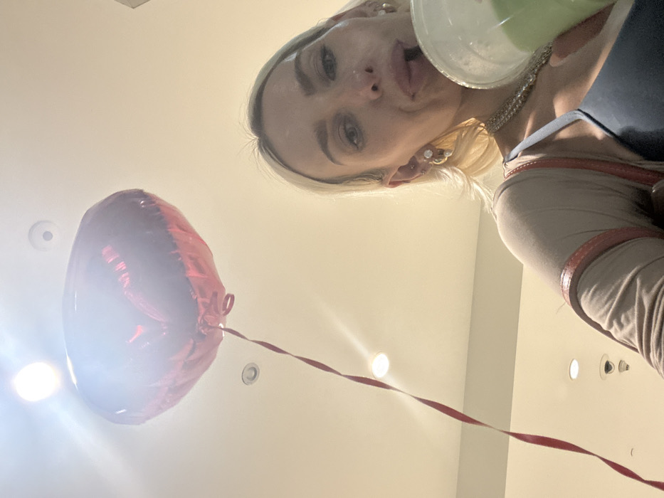From a low angle, a woman with blonde hair and silver jewelry holds a drink to her lips. A red balloon floats below her, attached to a long ribbon. The background is a light, neutral color, likely a ceiling, with several recessed lights. The woman is wearing a grey top with a reddish-brown trim. Her expression is playful, with her tongue slightly protruding.