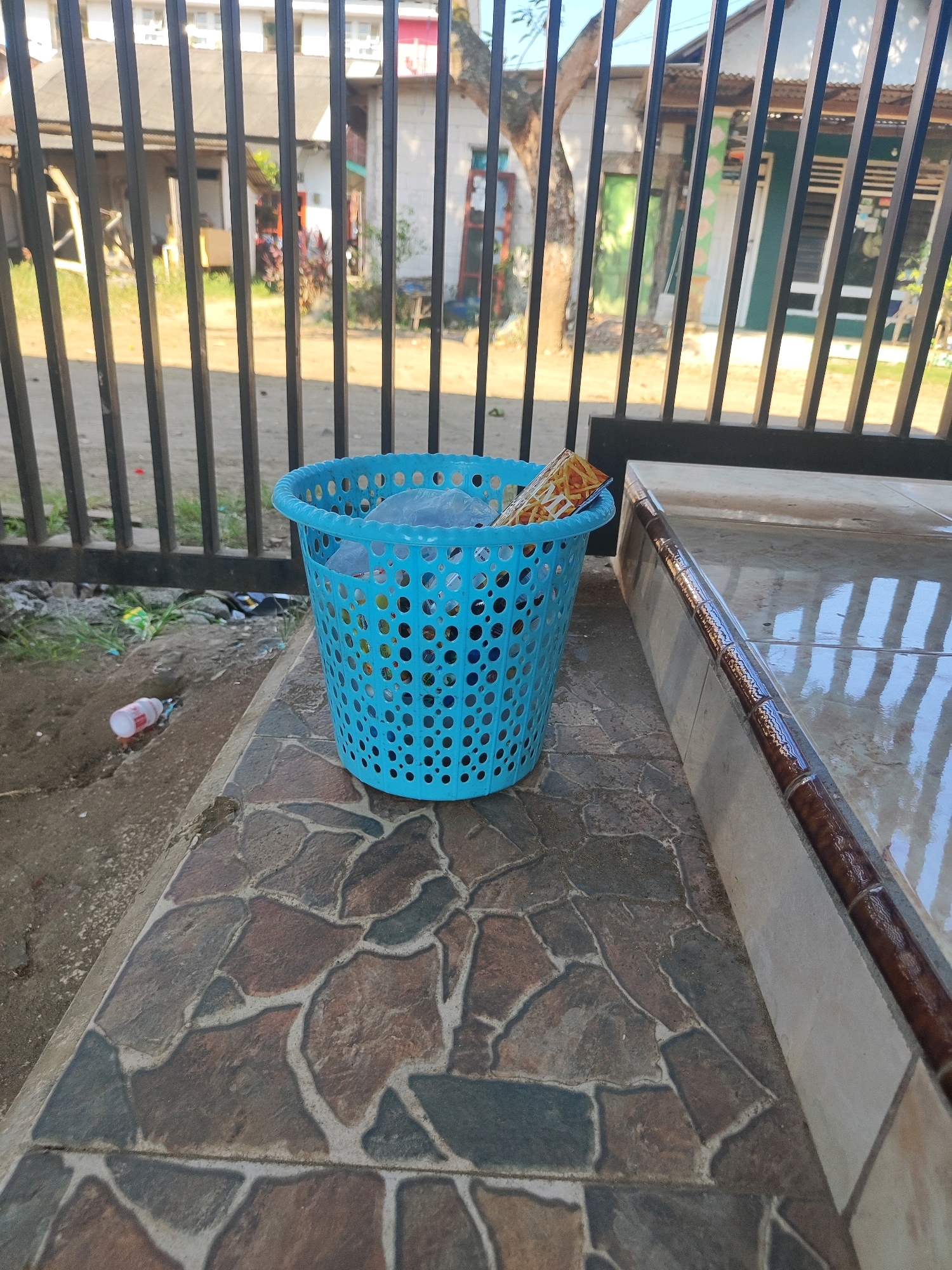 A blue plastic laundry basket is filled with trash, including plastic bags and a snack wrapper, and sits on a tiled patio in front of a black metal fence. To the left of the basket, a plastic bottle lies on the ground. In the background, a building with white walls and a red roof is visible. The scene is located in Tasikmadu, Indonesia.