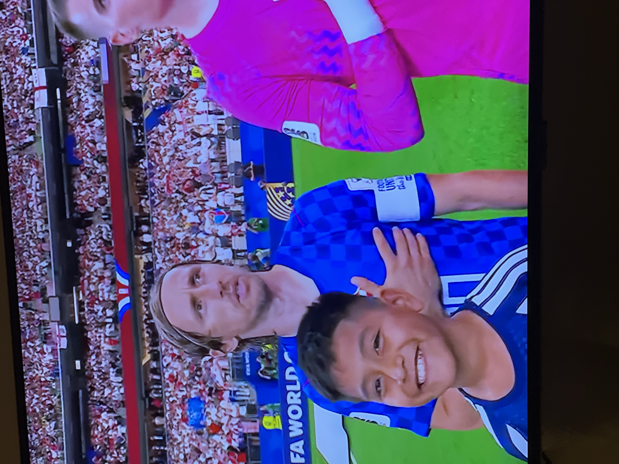 The image shows a close-up of a soccer match with a large crowd in the background. In the foreground, a young boy with dark hair and a wide smile stands in front of two soccer players. The boy is wearing a dark blue jersey with white stripes on the shoulders. The player on the left is wearing a blue and white checkered jersey, and the player on the right is wearing a bright pink jersey. Both players have their hands on their chests, presumably during a national anthem. The background shows a stadium filled with spectators wearing white and red attire, with some flags visible. The overall setting is a soccer field with a large audience. The image does not provide any information about the city of Florești, Romania.