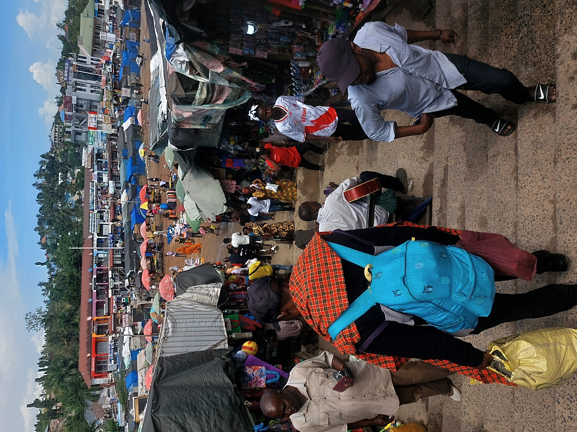 This bustling image captures a vibrant street market scene in Dar es-Salaam, Tanzania. The viewpoint is elevated, looking down onto a sloping paved area and a street lined with market stalls.
Numerous people populate the scene. In the foreground, a man wearing a light blue shirt, dark cap, and dark pants with Nike slides walks along the paved path, headphones visible. Below him, another man in a white shirt plays a guitar. A person dressed in a black top, red and black plaid wrap, and black pants wears a bright blue backpack adorned with cartoon characters. Near them, a yellow bag rests on the ground. Further down, a bald man in a khaki shirt holds a red-cased phone and another small object. Other individuals are seen throughout the market, including one in a white and red striped jersey (possibly "FIEBET" or "FIABET" with number "19").
The market stalls are characterized by various tarpaulins and umbrellas in blue, red, and yellow, providing shade for vendors and their merchandise. Goods like clothes, bags, and small items are displayed. A basketball and a small yellow ball are visible on one stall.
In the background, a hilly landscape is dotted with buildings and lush green tropical trees, including palm trees. Several buildings have visible text, including "THE AMAZON COLLEGE", "ASANAGI", "PATA SIMU MPYA" (Get a new phone), "VODACOM", "M-PESA", "SIMU" (Phone), and "CHAKULA HARAKA" (Fast Food).
The scene unfolds under a clear sky with scattered clouds, indicating daytime and fair weather. The overall impression is one of active daily commerce and community life in an urban setting.