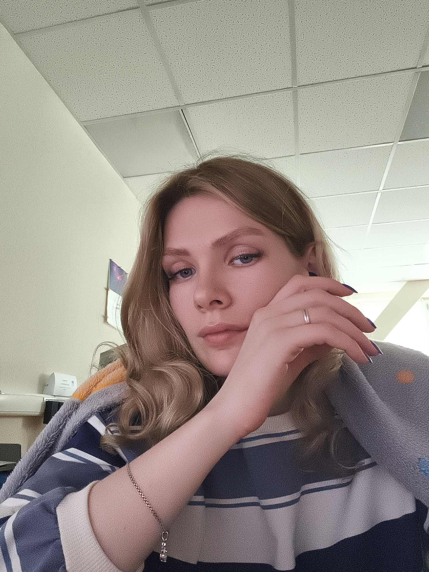A young woman with blonde wavy hair is sitting indoors. She is wearing a striped sweater and has a blanket draped over her shoulders. Her left hand is raised to her face, and she has a ring on her ring finger. Her right arm is visible and has a bracelet. The room appears to be an office or a similar institutional setting, with a ceiling with square panels and a wall in the background. The woman's gaze is directed towards the camera. The city of Postol, Russia is not identifiable as a feature of this image.