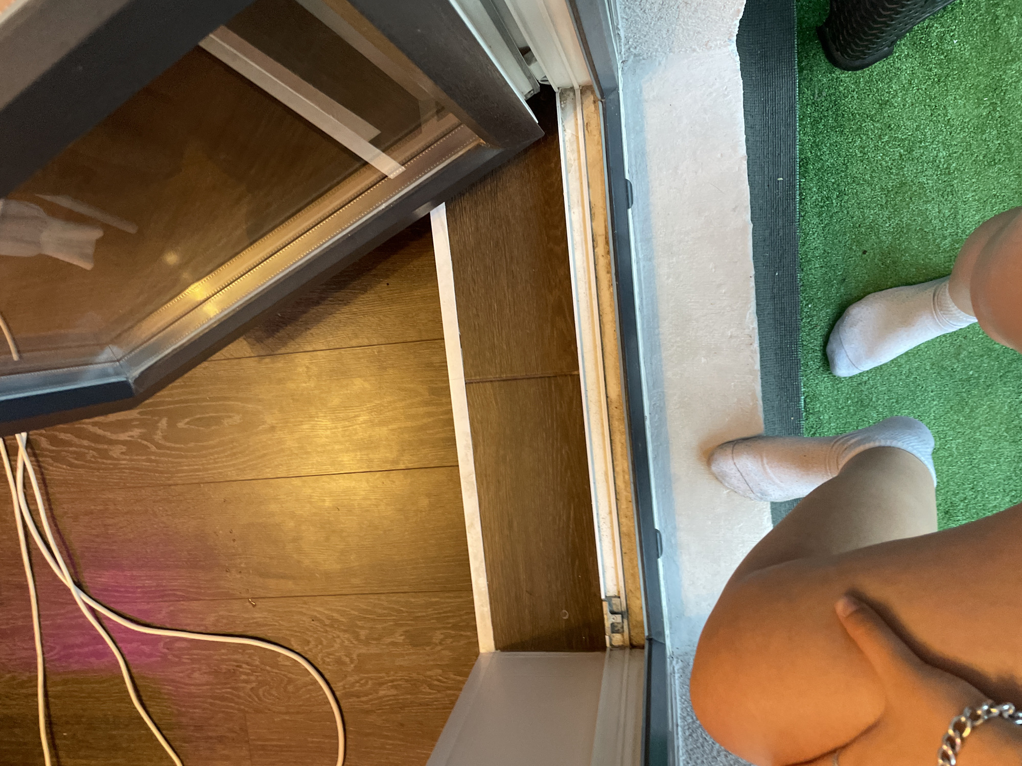 The image captures a close-up view of a person's legs and feet, wearing white ankle socks. The person is seated, with their left hand resting on their left thigh, adorned with a silver bracelet. The scene is set indoors, near a door or window, with a wooden floor visible. A white electrical cord snakes across the floor, partially illuminated by colored lights. The immediate area around the door appears to be a transition space, with a strip of darker flooring leading to a green, artificial turf-like surface. In the background, a partially open window or door reveals a reflection on its glass. The environment suggests a domestic setting. No specific Sinyavino, Russia location details are identifiable in the image.