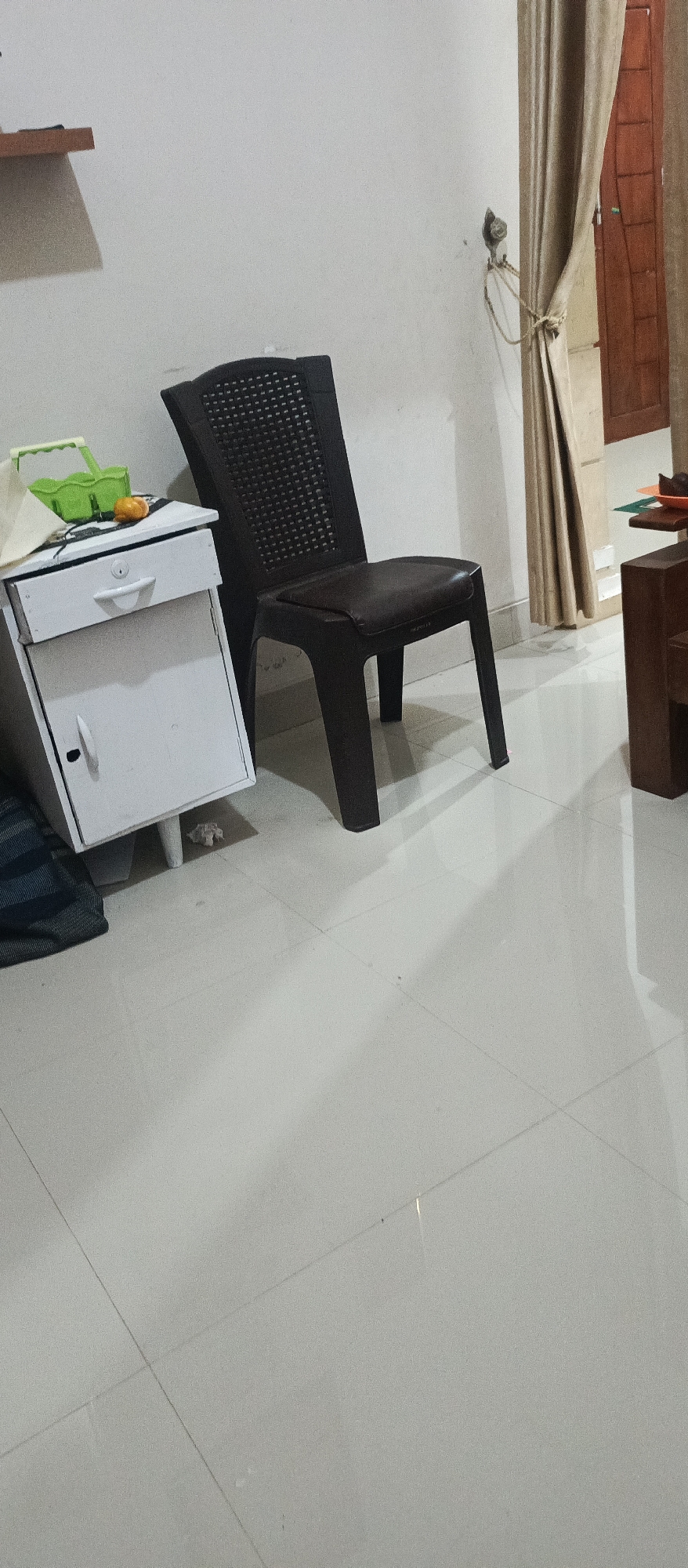 The image shows an indoor scene with tiled flooring. On the left, there is a small white cabinet with a drawer and a door. A green plastic basket and an orange object are on top of the drawer. To the right of the cabinet is a dark brown plastic chair with a woven seat and back. In the background, a light brown curtain is drawn to the right, revealing a portion of a wooden door. A small shelf is mounted on the wall above the cabinet. The overall impression is of a simple, functional indoor space. This scene is consistent with a residential or office interior found in Arjasa, Indonesia.