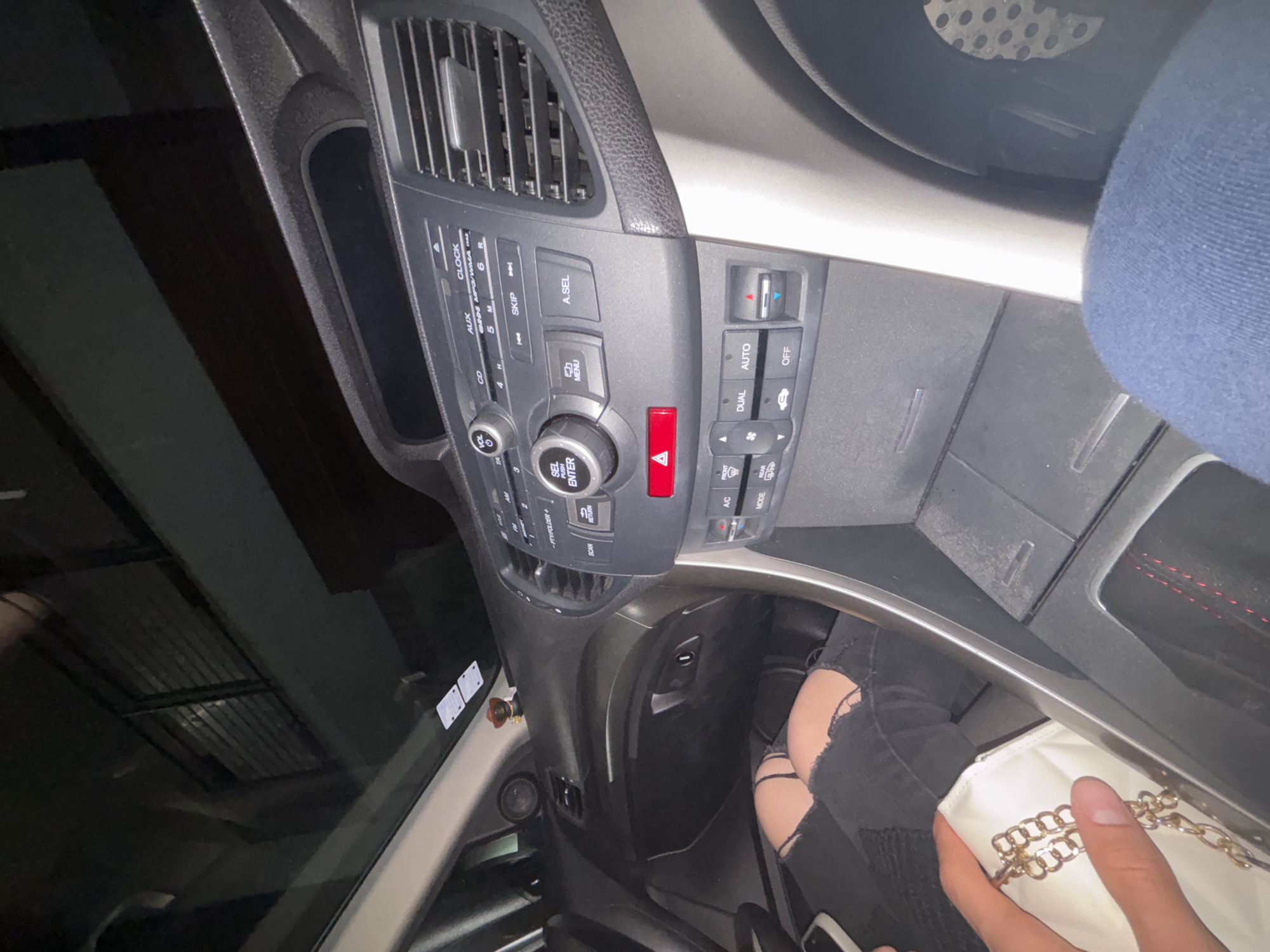 The image shows the interior of a car. The dashboard is visible, featuring the car's infotainment system and climate control panel. A person wearing ripped black jeans is holding a white purse with a gold chain. The location is impossible to determine without more context or information.