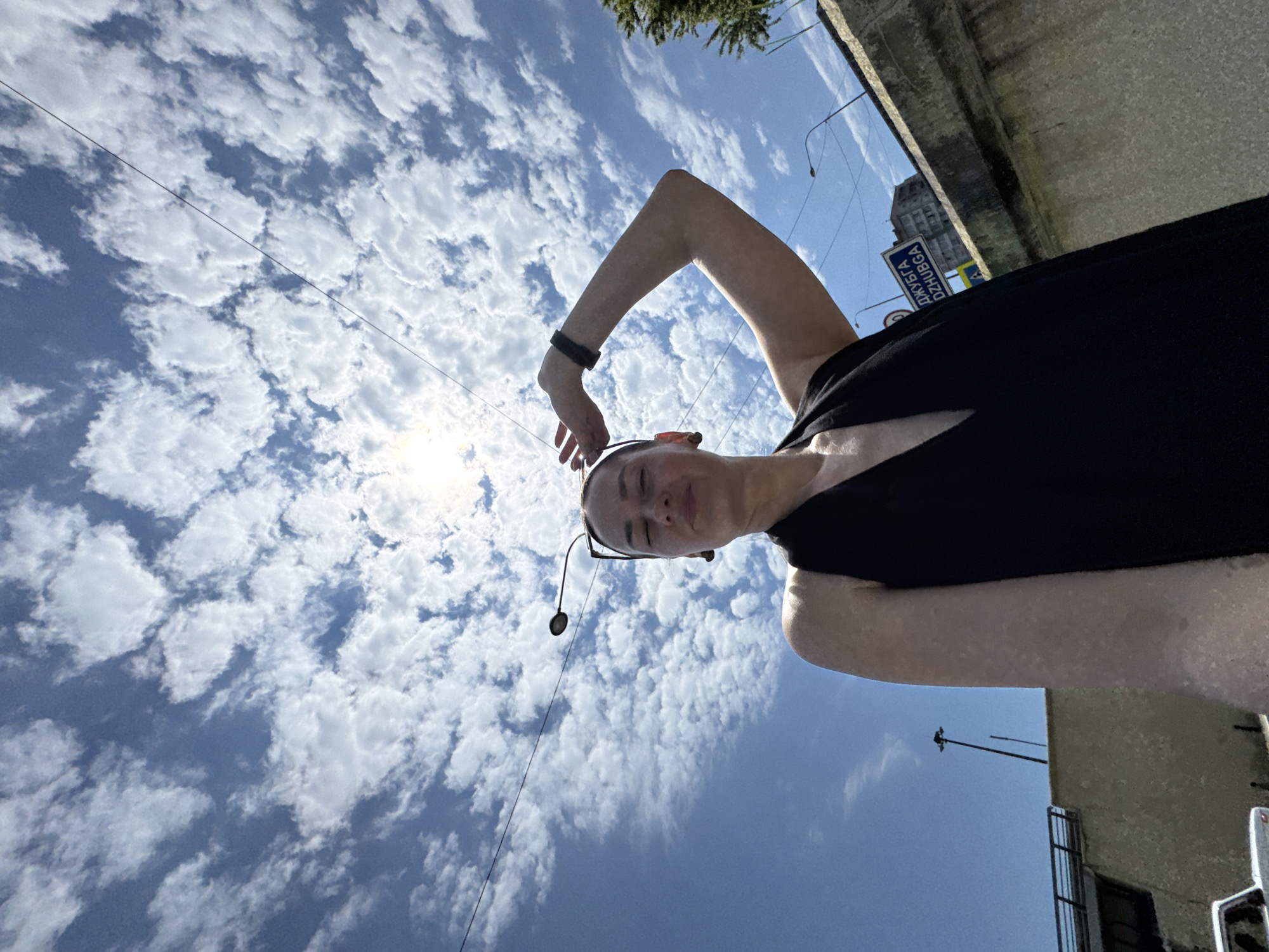 The image is a wide-angle, low-angle selfie taken outdoors on a sunny day with a blue sky and scattered white clouds. The sun is bright and positioned high in the frame. A woman is the central subject, wearing a black sleeveless V-neck dress and a black wristband. She is looking upwards with her eyes closed and one hand raised to her forehead. Her arm is extended, casting a shadow.
In the background, power lines crisscross the sky. A street lamp on a pole is visible to the left of the woman. A concrete barrier runs horizontally across the lower right portion of the image. A blue street sign with white text, which reads "ДЖУБГА DZHUGBА", is attached to the barrier. Behind the barrier, there is a building and a tree. The presence of the street sign indicating "Dzhubga" strongly suggests the location is near or within the Dzhubga settlement, which is a resort area within the Greater Sochi region of Krasnodar Krai, Russia. A partial view of a white car is visible in the lower left corner.