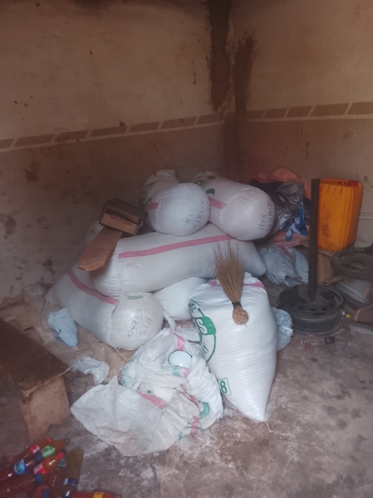 The image depicts a cluttered indoor space in Gwong, Nigeria. The room has aged walls with a visible tile border. Numerous white sacks are piled in the center, several with the inscription "HIS GRACE." A wooden block rests atop the sacks. A broom leans against one of the sacks, and there is an orange plastic container and a black wheel rim in the background. On the lower left, several plastic bottles are scattered on the floor. The floor itself appears to be bare and stained. No people are visible in the image.