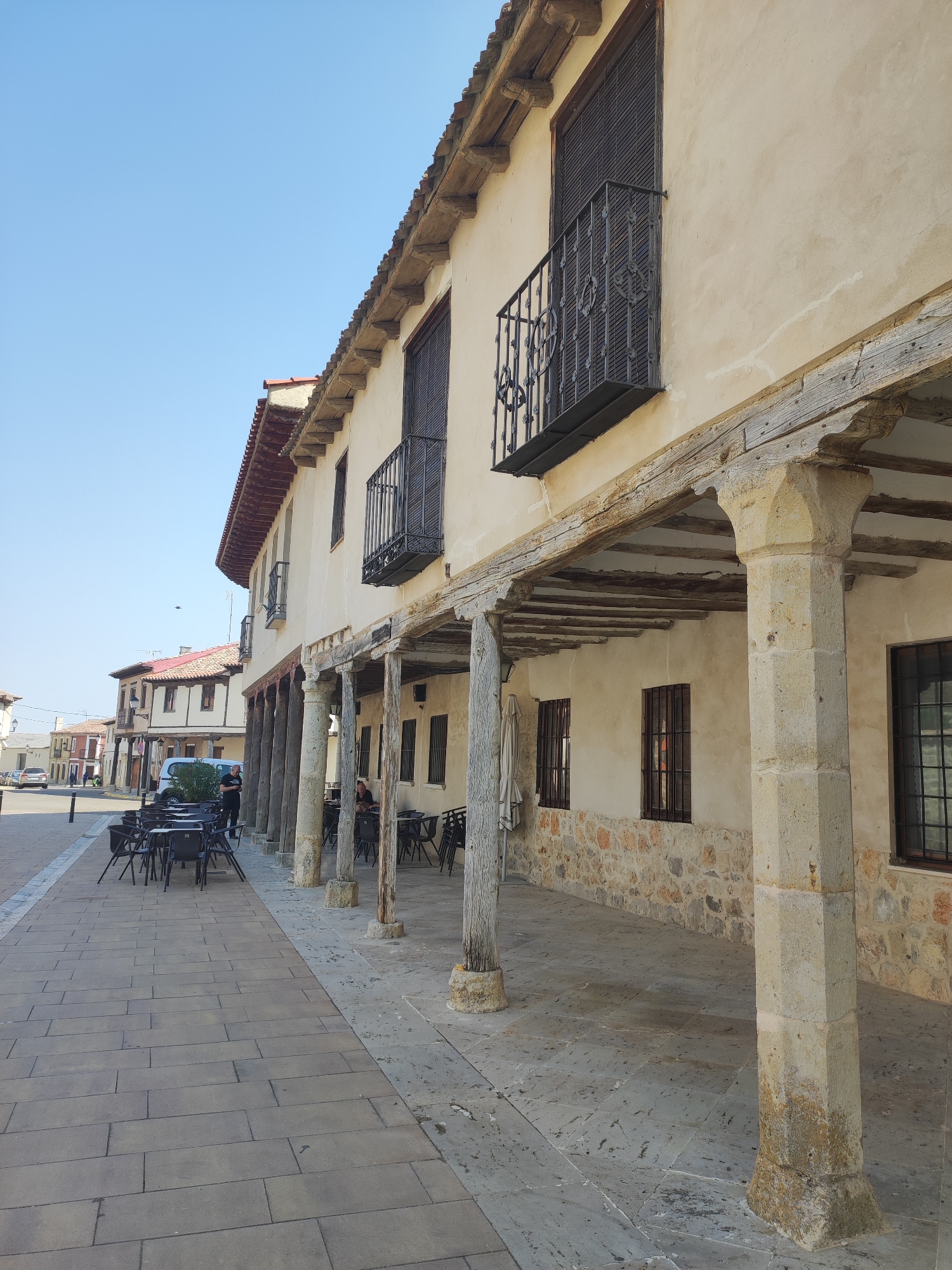 The image captures a street scene in Ampudia, Spain, dominated by the facade of a two-story building with a covered walkway. The building features a light-colored stucco exterior, with a textured stone base on the ground floor. Dark metal balconies with ornate railings adorn the upper level. The covered walkway is supported by a series of large, rough-hewn stone pillars and heavy wooden beams, showcasing a rustic architectural style. Several tables and chairs, typical of outdoor seating for a cafe or restaurant, are arranged under the walkway and on the tiled sidewalk. Beyond this building, other structures with red-tiled roofs and traditional Spanish architecture are visible down the street. The sky is clear and blue.