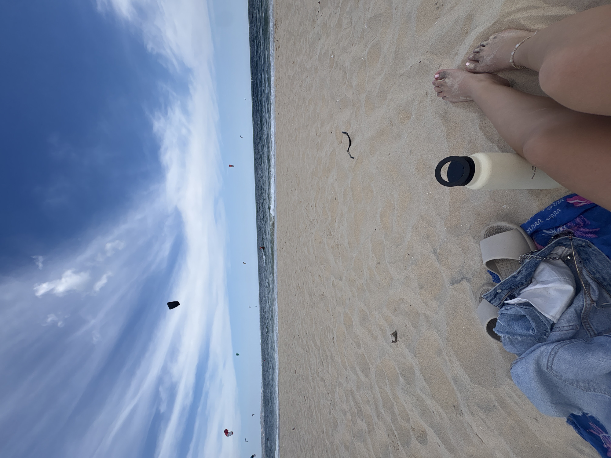 A first-person perspective shot captures a sandy beach with the ocean and sky in the background. In the foreground, a person's bare legs and feet are visible, adorned with a delicate anklet. To the left, a light-colored water bottle with a black lid rests on the sand. Beside it, a pair of light-colored slides and a denim jacket are casually placed. The beach is a light tan color with visible sand ripples. In the middle ground, the dark blue ocean meets the shore, with gentle waves rolling in. The sky is a vibrant blue, streaked with wispy white clouds. Several colorful kites are scattered across the sky, suggesting a windy day. The scene is set in the city of Phan Thiết, Vietnam, a popular destination known for its beaches and water sports.