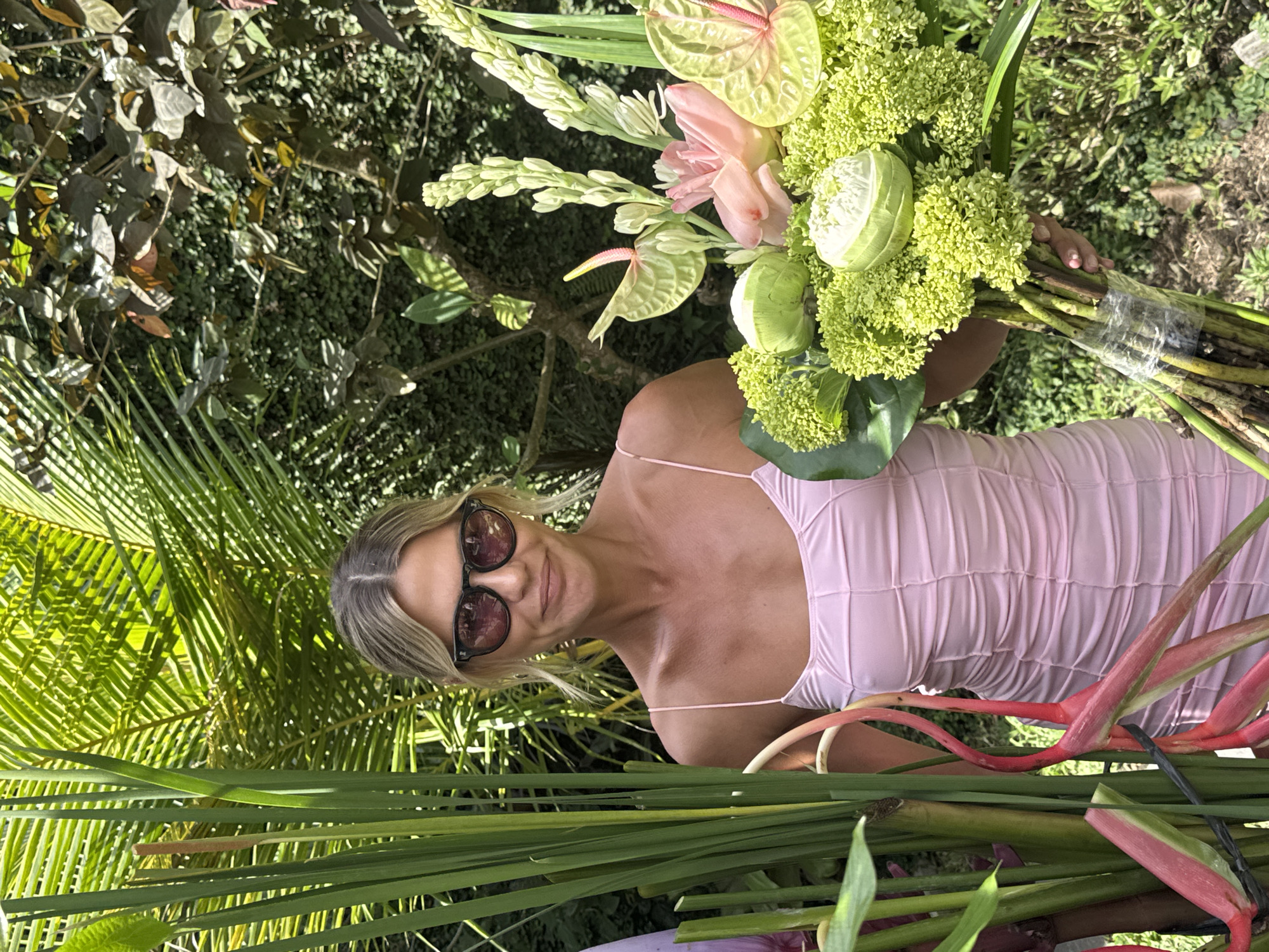 A woman with blonde hair and sunglasses is holding a large bouquet of tropical flowers and foliage. She is wearing a light pink dress. The setting appears to be outdoors in a lush, green environment, likely in North Kuta, Indonesia, indicated by the tropical flora present, like heliconia, anthuriums, and palm fronds.