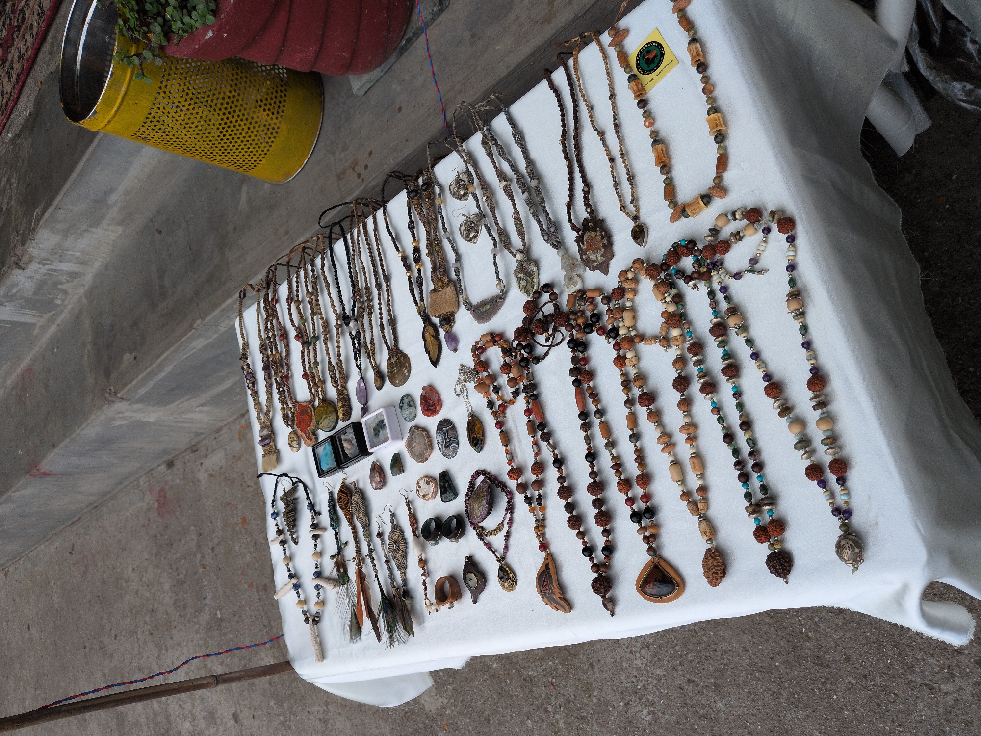 A stall displays a collection of handmade jewelry, primarily necklaces, laid out on a white cloth covering a table. The jewelry features a variety of beads made from natural materials like wood, seeds, and stones, interspersed with polished stones, and some pieces with feathers and metal charms. Several necklaces have pendants, including a large teardrop-shaped one with a wooden frame and a dark brown stone, and another with a carved wooden leaf. Smaller items like rings and earrings are also present. In the background, concrete steps rise, and a yellow perforated trash can and a red planter are visible, suggesting an outdoor market or street setting in Haini, India. The ground is paved with concrete.