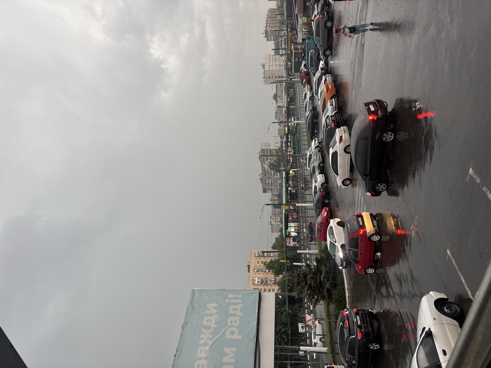 The image shows a rainy day in Kharkiv, Ukraine, overlooking a parking lot. Cars are parked in rows, reflecting the rain on the asphalt surface. In the distance, buildings define the cityscape. A person with an umbrella is seen walking among the parked vehicles. A large sign with Cyrillic text is visible in the foreground.