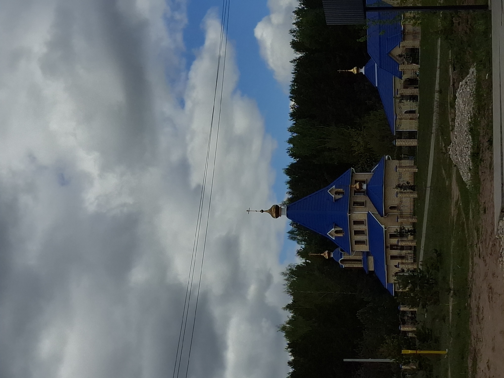 The image depicts a building in Chaikovsky, Russia. The building has a blue roof with gold accents and is surrounded by trees. The sky is cloudy with some blue patches. There are power lines visible in the sky. The building appears to be a church or religious building. There is a dirt path in the foreground.