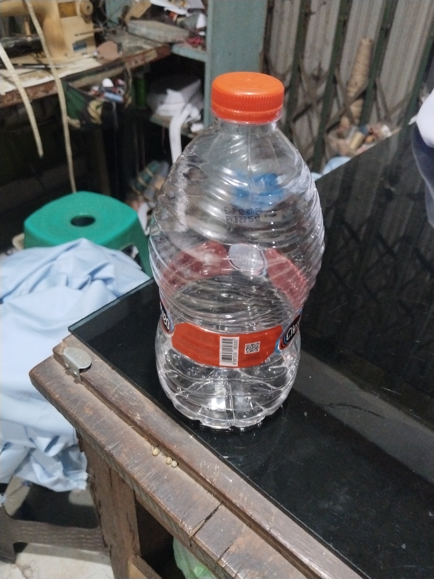 An eye-level, medium shot captures a clear plastic water bottle with an orange cap standing upright on a dark, reflective surface, likely a table. The bottle is partially filled with water, and its label is red with some visible text and a QR code. The background is blurred, suggesting an indoor environment. To the left of the bottle, a folded light blue cloth is visible, and further back, a green plastic stool can be seen. In the upper left quadrant, partially obscured by the bottle, is a sewing machine on a workbench, surrounded by various tools and materials. The overall setting appears to be a workshop or a domestic area where crafting or repairs might take place. The lighting is moderate, with some reflections on the table surface. There are no people present in the image. The scene does not provide any specific geographical markers to indicate a location within the city of Unknown, Indonesia.