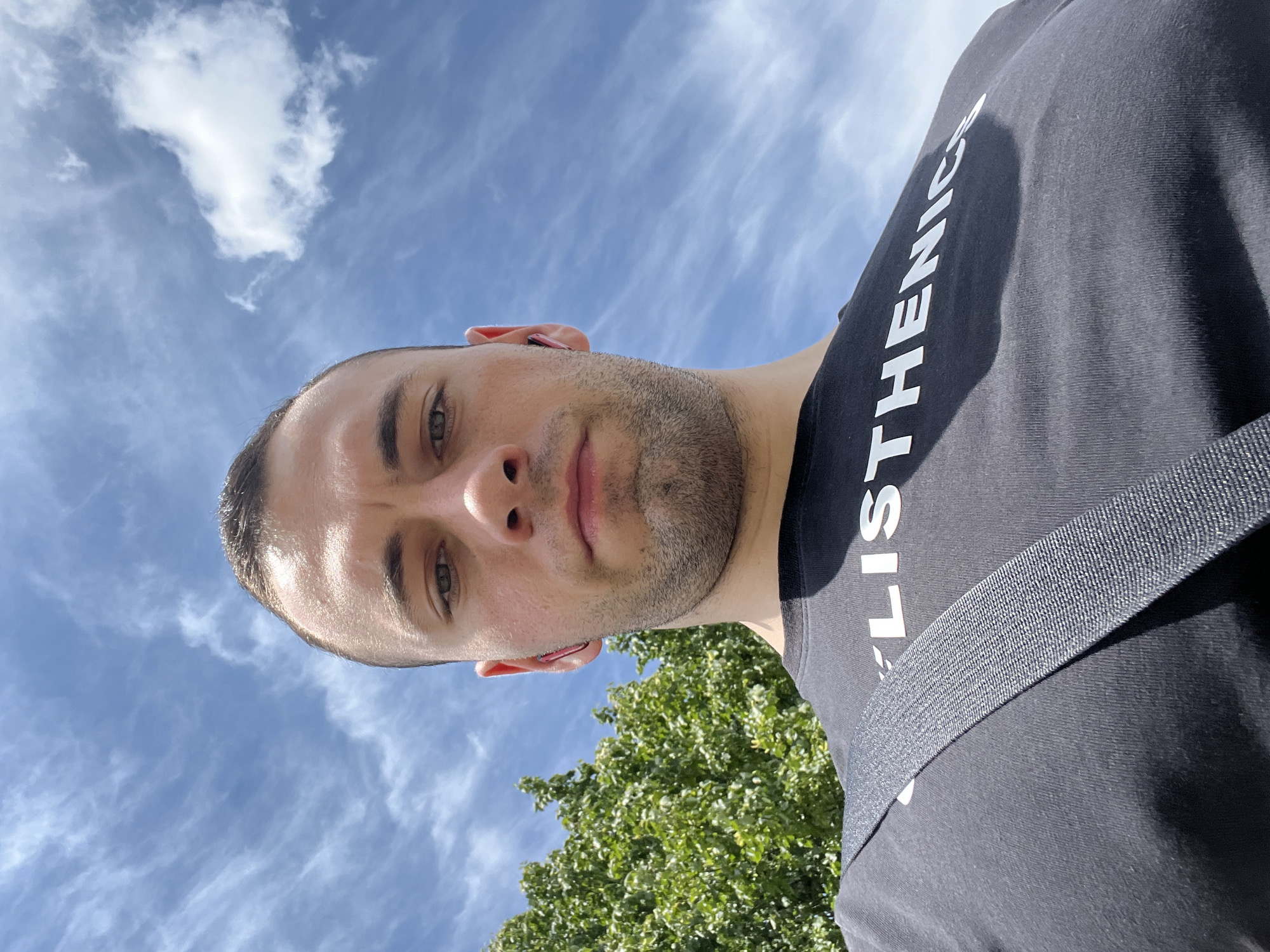 The media file displays a close-up, low-angle shot of a man's head and upper torso, rotated 90 degrees clockwise. The man, with short hair and light stubble, is looking directly at the camera with a neutral to slightly serious expression. His eyes appear to be light-colored, possibly green or hazel. A small red earbud is visible in his right ear. He is wearing a black t-shirt with white text partially visible on the right side, reading "LISTHENICS" (likely part of "CALISTHENICS"). A dark grey strap, possibly from a bag, crosses his body. The background features a clear blue sky with white, wispy clouds, indicating bright daylight and sunny weather. Green tree foliage is visible in the lower right portion of the frame, suggesting an outdoor setting in a natural or urban green space.