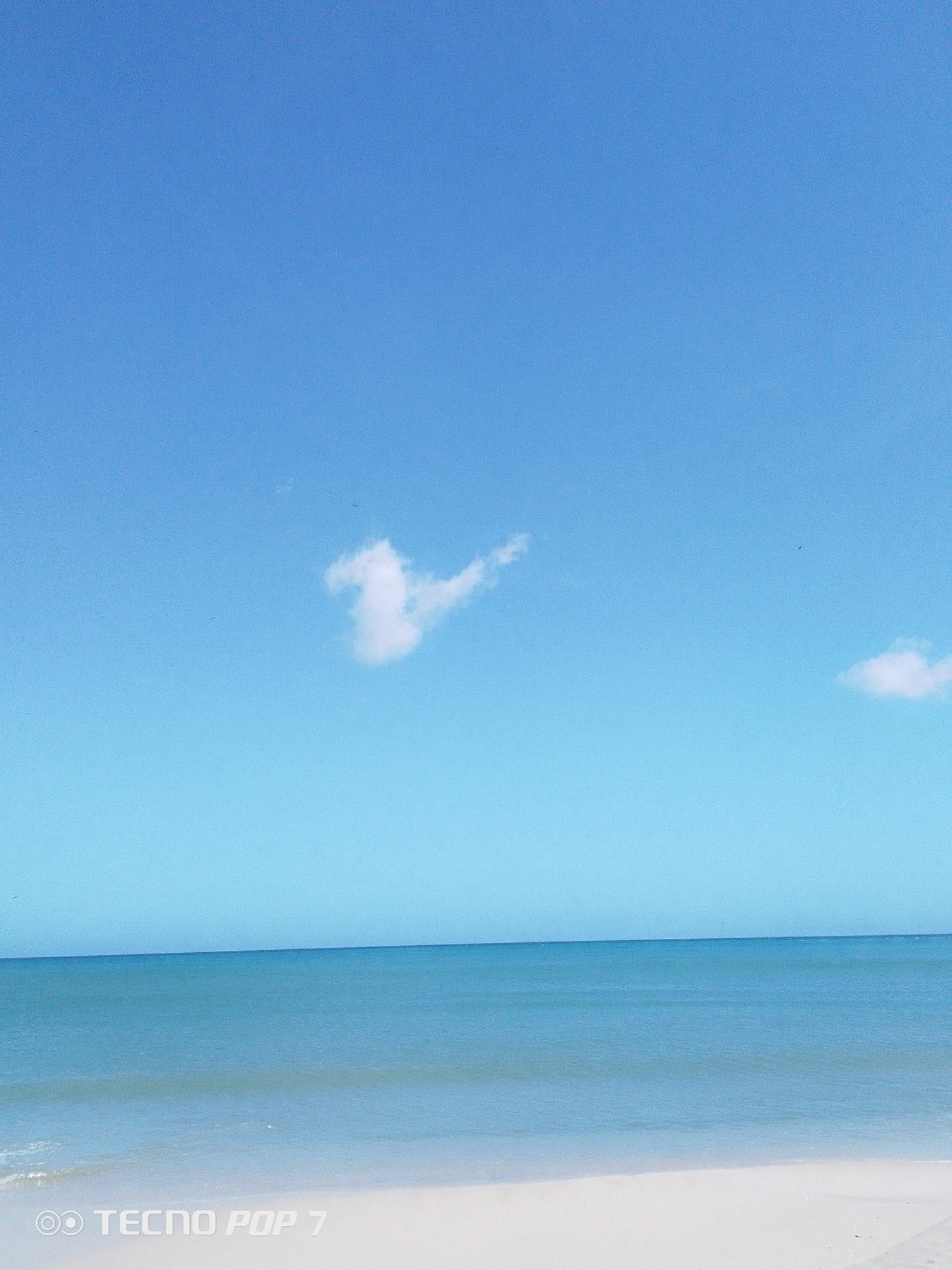 The image shows a tranquil beach scene. The sky is predominantly blue with a few scattered white clouds. The ocean is calm, with a gradient of turquoise hues. The beach sand is light-colored. The location is Pedro González, Venezuela. There are no people visible in the image. The focus is on the natural elements.