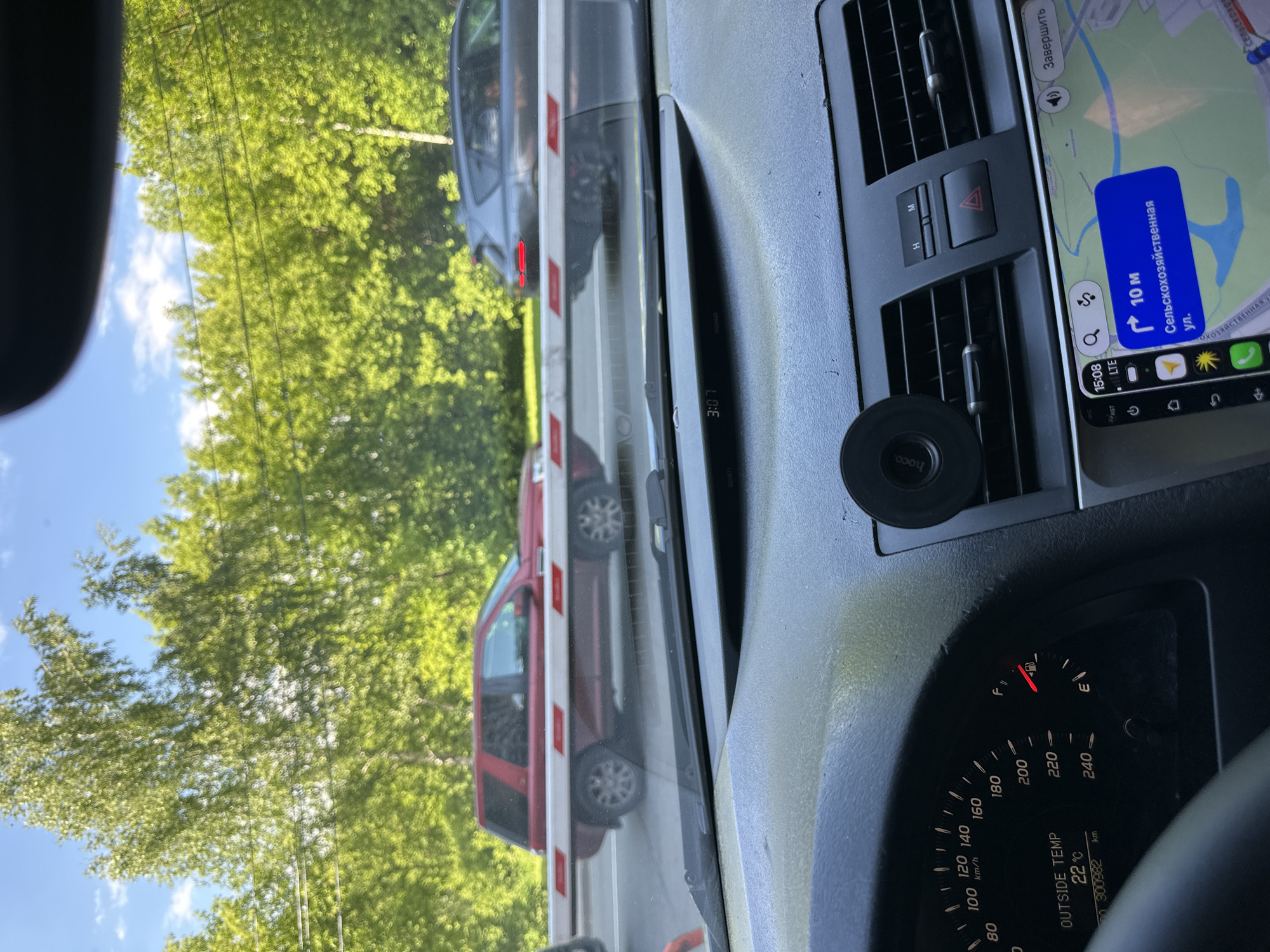 The image captures a first-person perspective from inside a car, looking out through the windshield at a stopped vehicle and a barrier. The scene suggests the car is waiting at a gate or railway crossing.
In the foreground, the car's interior is visible, including air vents, a hazard light button, and a phone holder branded "hoco.". A smartphone is mounted to the right of the dashboard, displaying a navigation app. The app shows "15:08" as the time, "LTE" signal, and a turn instruction: "10 M" for "Сельскохозяйственная ул." (Selskokhozyaystvennaya St.), confirming the location in Moscow, Russia. Another digital display shows "3:07". Part of the car's instrument cluster is visible, showing "OUTSIDE TEMP 22°C", an odometer reading "300982 km", and a full fuel tank. The speed dial is also visible, marked in "km/h" up to "240".
Through the windshield, a white and red striped barrier is visible, indicating a stop. A dark grey car is stopped in front of the barrier, and a red car is further ahead. Lush green trees line both sides of the road, and a clear blue sky with some white clouds is overhead, suggesting a bright, warm day. The image is rotated 90 degrees clockwise.