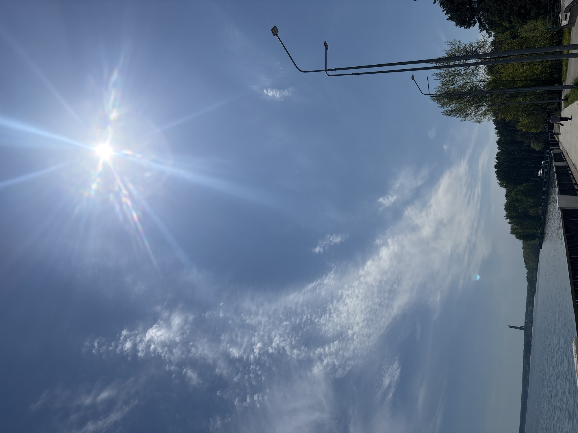 The image shows a bright, sunny sky over a body of water, likely a lake or river, located in Chaikovsky, Russia. A paved walkway runs along the edge of the water, lined with trees and streetlights. A person is walking along the path. The sky is blue with scattered cirrus clouds and a bright sun creating lens flares. The water is calm and reflects the sky.
