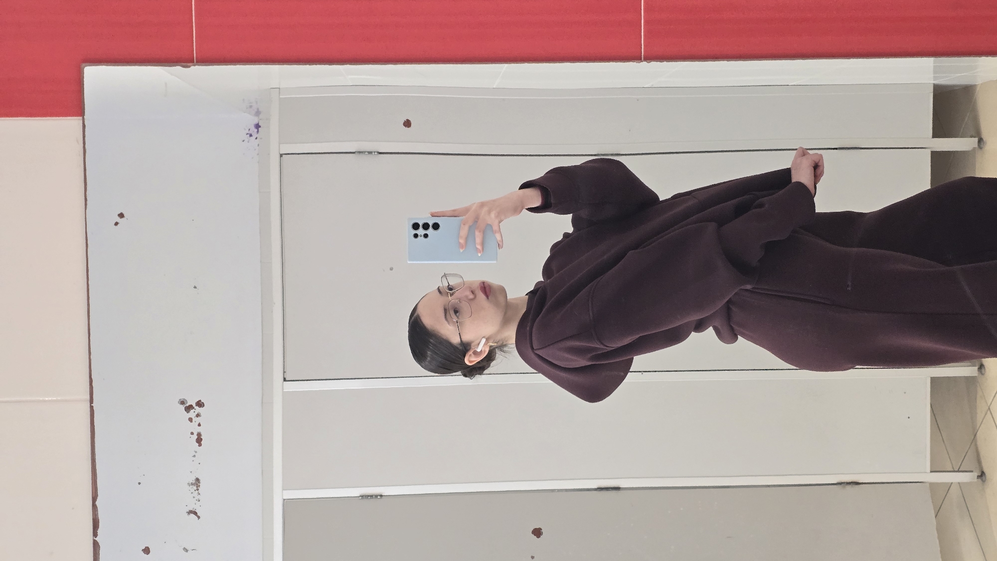 A person is reflected in a mirror, taking a selfie with a light blue smartphone. The individual has dark hair pulled back into a bun, wears glasses, and has a white wireless earbud in one ear. They are wearing a maroon or dark purple hooded sweatshirt or coat.
The reflection shows a restroom stall with a red and white tiled wall at the top of the frame. There are white panels that make up the stall's walls, which have a few marks and stains. The stall appears to be located in Stara Zagora, Bulgaria, based on the provided context.