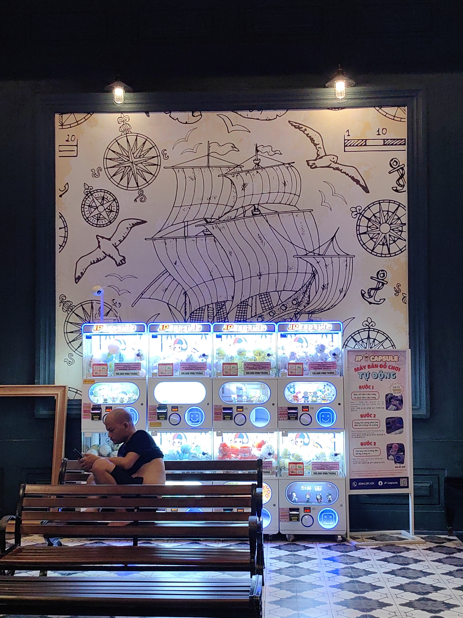 An indoor scene in Bai Chay Ward, Vietnam, features a man seated on a wooden bench, engrossed in his phone. Behind him is a row of several brightly lit "JP Capsule" vending machines filled with various toys. Above the machines and encompassing the upper wall is a large, hand-drawn mural with a nautical theme.
The mural depicts a large sailing ship with multiple sails, surrounded by stylized compass roses, anchors, and flying seagulls. Two small, cage-like light fixtures are mounted near the top edge of this artwork, casting a warm glow downwards.
The vending machines are uniform in design, displaying glowing circular buttons and screens. Text on the machines indicates "100.000 VNĐ/TRỨNG" and "150.000 VNĐ/TRỨNG," suggesting the price per capsule. To the right of the machines, a large white sign titled "JP CAPSULE MÁY BÁN ĐỒ CHƠI TỰ ĐỘNG" provides instructions in Vietnamese, including "BƯỚC 1: Đọc số tiền trên máy và đưa tiền vào máy," "BƯỚC 2: Quay một sang phải cho đến khi trứng rơi xuống," and "BƯỚC 3: Cho tay vào ở sáng đèn để lấy trứng ra." It also lists a phone number, "0344.391.431," and the brand "JP Capsule," alongside "ĐẾ TỪ 100k" and "NƠI LẤY QUẢ." The floor is tiled with a repeating geometric pattern in shades of blue and white. The man appears focused on his device, creating a casual and modern moment within a space designed with a whimsical, adventurous aesthetic.