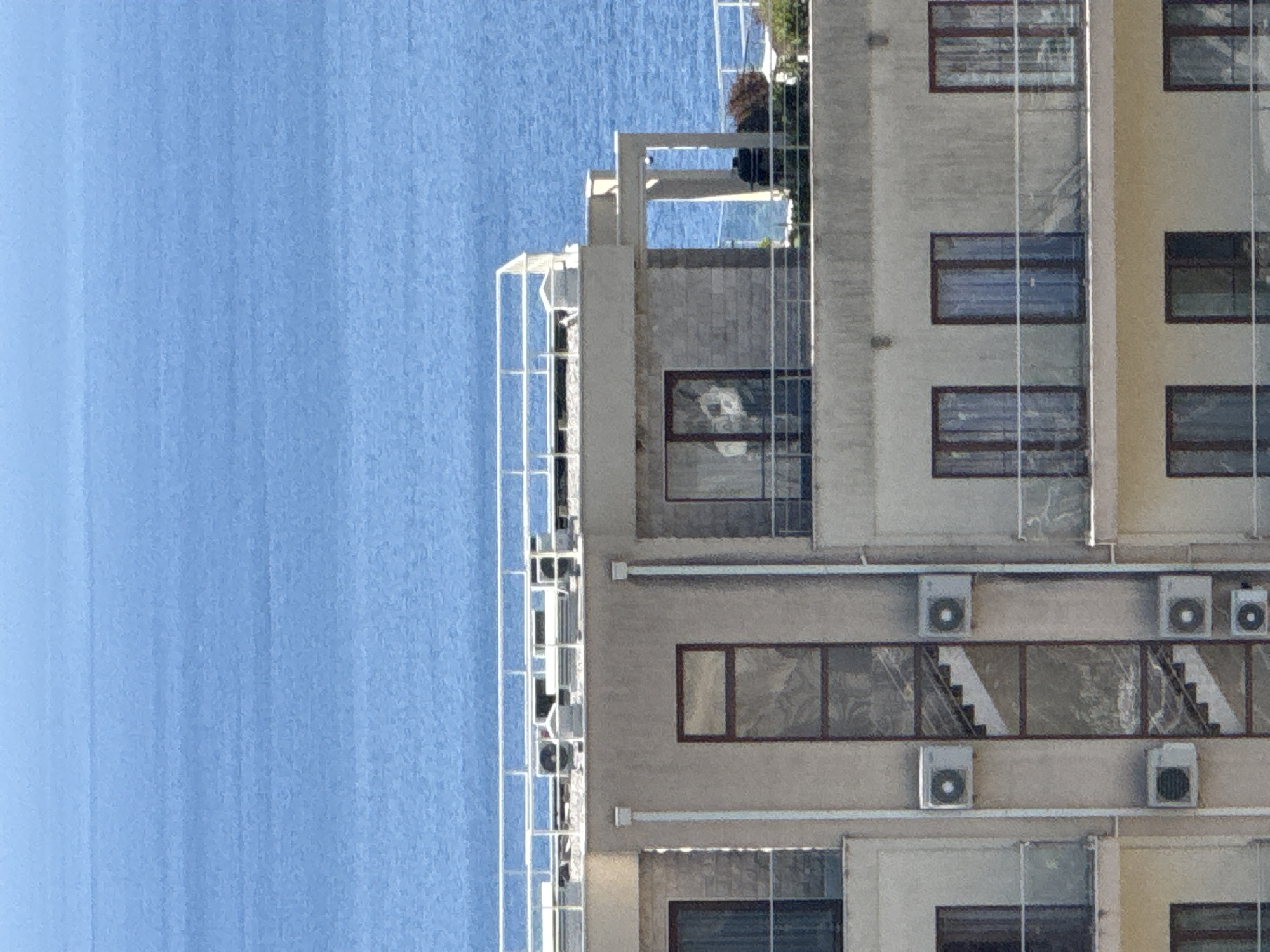 The image shows a close-up view of the upper floors of a beige apartment building with a large body of blue water in the background, likely the Adriatic Sea. The building features multiple balconies with glass railings, and several windows with dark frames. On the facade of the building, there are multiple white air conditioning units mounted on the walls. In the center of the building, a vertical section with glass panels reveals a staircase inside. The rooftop area of the building has white railings and some mechanical equipment, possibly air conditioning units or vents. The water in the background appears calm with slight ripples. The scene is likely located in a coastal area of Boreti, Montenegro, given the architecture and the prominent presence of the sea.