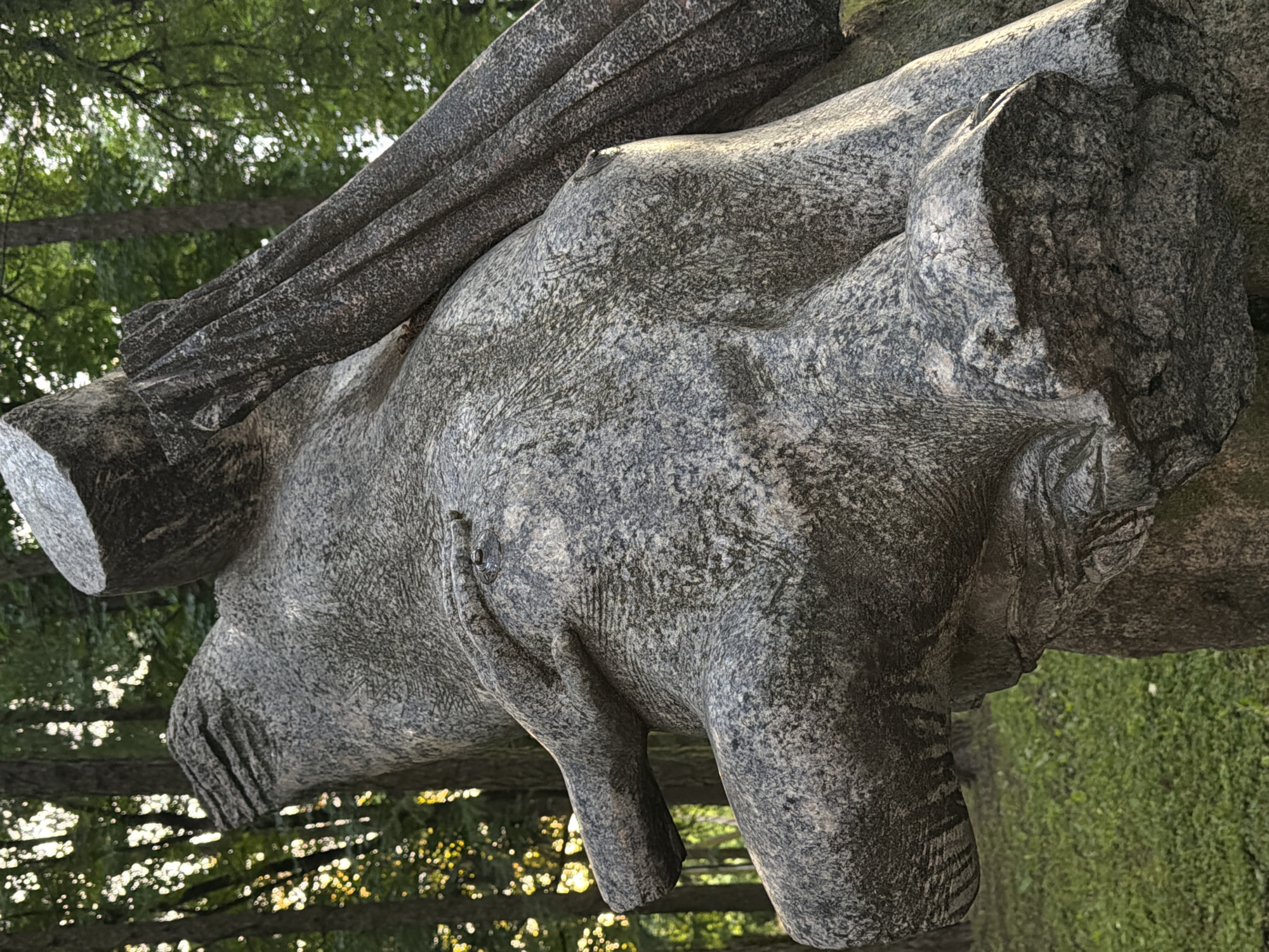 The image shows a close-up view of a large, rough-hewn granite sculpture outdoors, possibly in a park or garden. The sculpture appears abstract, with some parts resembling a body or limbs, and another section with textured, draped folds. Sunlight filters through the trees in the background, creating a dappled effect on the sculpture and the green grass and foliage. The sculpture's texture is granular and varied, with shades of gray and black. A hand-like appendage is visible touching a rounded, torso-like section of the sculpture. The overall impression is of a weathered, perhaps ancient-looking piece of art set against a natural backdrop. This sculpture is located in the Serebryany Bor park in Solnechnogorsk, Russia.