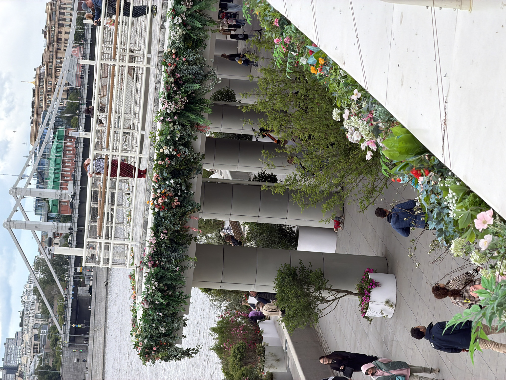 The image shows a partially-covered walkway, elaborately decorated with greenery and flowers. Below the walkway is a body of water, likely a river, and on the opposite bank are buildings typical of a city. People are visible both on the walkway and below it. The walkway is supported by cylindrical pillars. The location appears to be in Moscow, Russia, specifically near the Patriarshy Bridge.