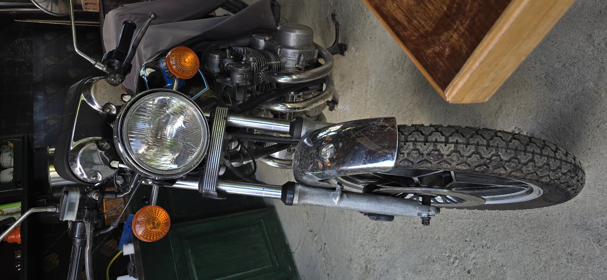 This is a close-up, overhead shot of the front of a motorcycle, taken from a slightly elevated angle and rotated 90 degrees clockwise. The motorcycle is stationary and appears to be in a garage or workshop, with a concrete floor and a partially visible dark green door in the background. To the right of the motorcycle, a wooden object with a warm, polished finish is visible.
The motorcycle's front end is the primary focus. A large, chrome-rimmed headlight is prominent, with a clear lens reflecting light. To the left and right of the headlight are orange, circular turn signals. The motorcycle's engine, with its metallic pipes and fins, is visible behind the headlight assembly. Chrome accents and detailing are visible on various parts of the bike, including the handlebars and parts of the front suspension. The front tire, with its distinct tread pattern, is also clearly visible.
There are no people present in the image. The lighting suggests it might be daytime, possibly indoors with artificial lighting. The overall impression is that of a detailed view of a well-maintained, classic-style motorcycle. No text is clearly legible within the image.
