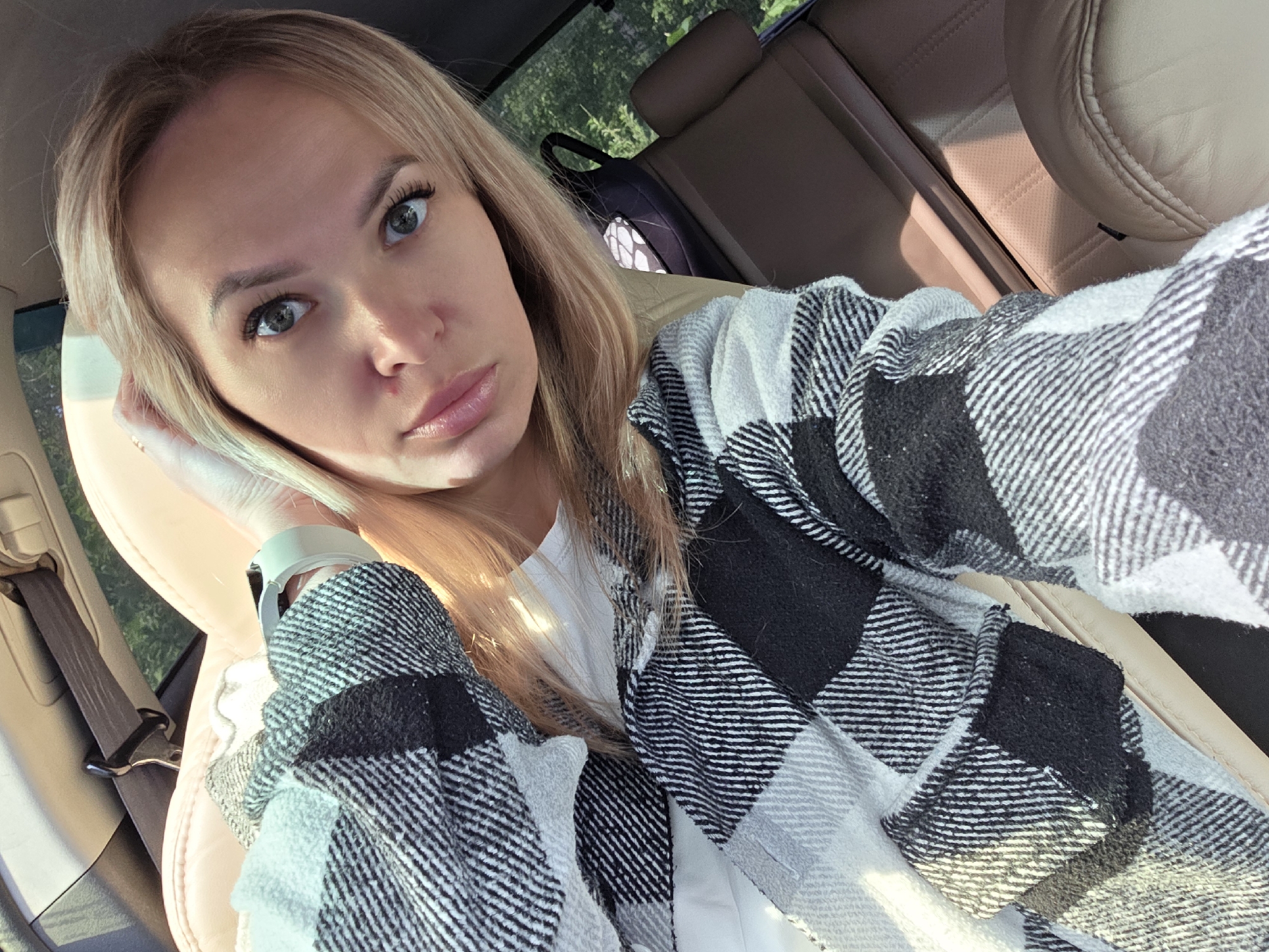 A young woman with blonde hair is taking a selfie from the driver's seat of a car. She is wearing a black and white plaid jacket over a white shirt and has her left hand on her head. Her eyes are open and she is looking at the camera. The car's interior is visible, including a tan leather seat and a seatbelt. Outside the car, trees are visible through the window. The lighting suggests it is daytime, possibly in Chaikovsky, Russia, given the context of the prompt.