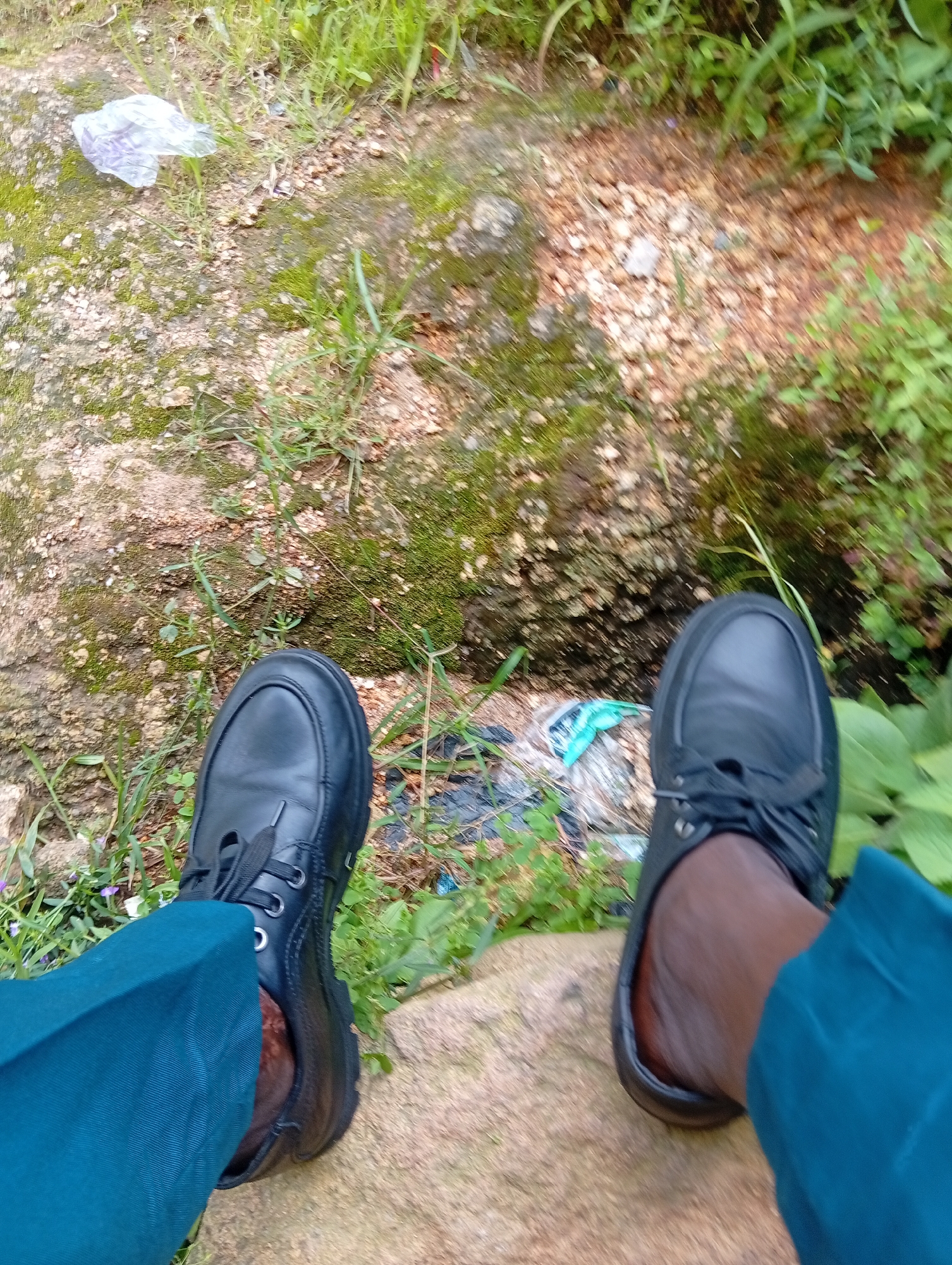 The image shows a close-up, overhead view of a person's legs and feet, wearing dark blue trousers and black leather shoes. The person is sitting on a large, rough, rock-like surface, which appears to be an outdoor environment. The ground is covered with small stones and patches of moss. Green vegetation, including grass and leaves, surrounds the area, suggesting a natural or semi-natural setting. In the background, some white and blue plastic debris is visible on the ground. The overall scene is consistent with an outdoor location in Kabong, Nigeria, likely a natural area or a less-developed part of the city where such terrain and vegetation are common.