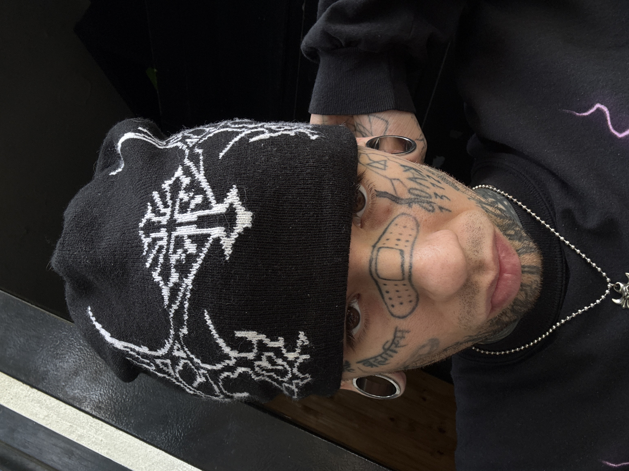 A person with extensive face tattoos wearing a black beanie with white patterned designs is depicted. The beanie covers the upper part of their forehead. Tattoos include a band-aid on the nose, text on the cheek, and geometric designs. The person has stretched earlobes with silver tunnels. They are wearing a black long-sleeved shirt with a lightning bolt design and a silver chain necklace. The background includes a black surface with a silver/white line. The orientation of the photo is rotated. There is no indication of Buenos Aires, Argentina.