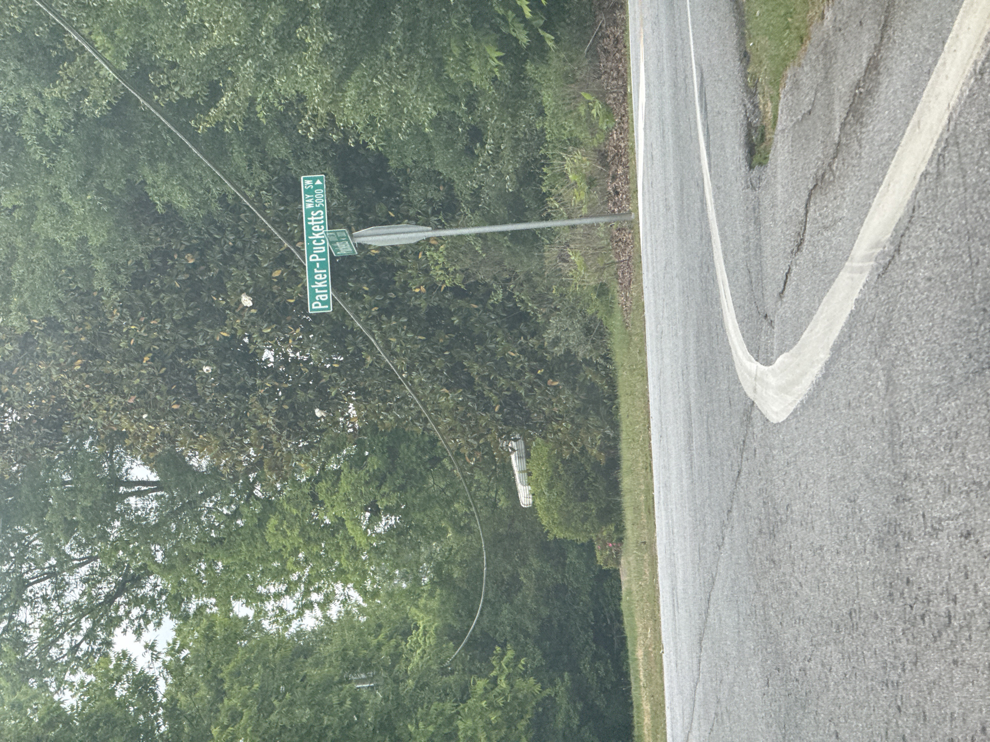 A daytime street scene shows a road sign for "Parker-Pucketts WAY SW 5000," which indicates the location of Parker-Pucketts Way Southwest, number 5000. The sign is green with white lettering and includes a directional arrow. Adjacent to the sign is dense green foliage from trees and undergrowth. The road is gray asphalt with a white lane marker. The location is in Mountain Park, United States of America.
