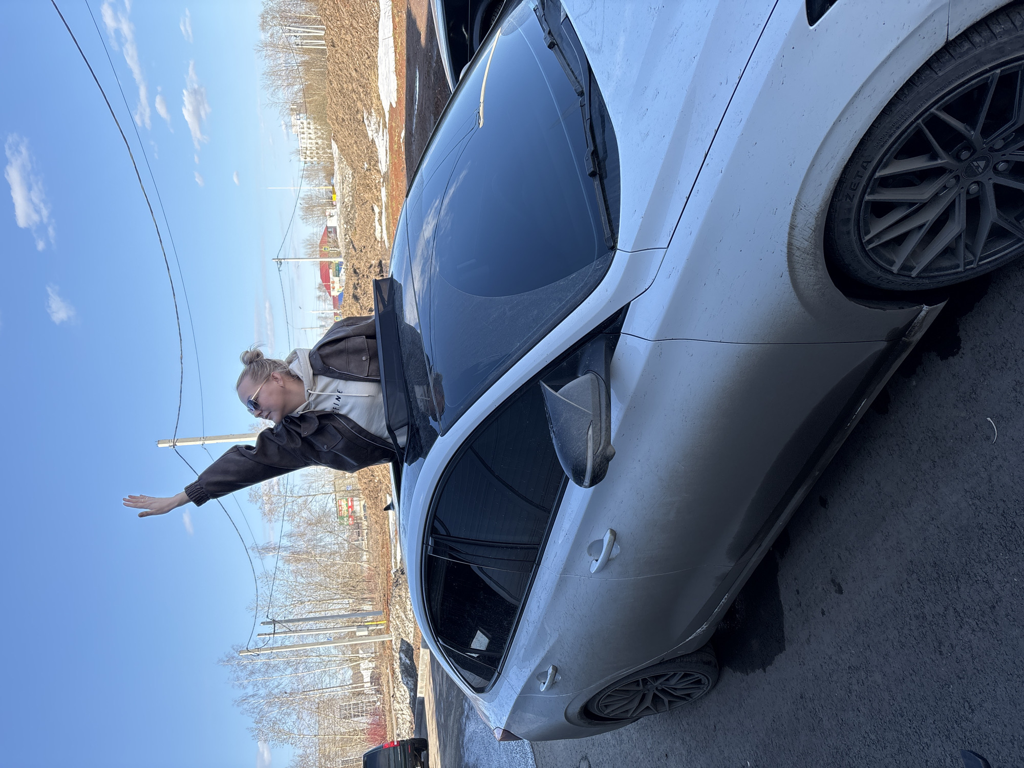A woman with blonde hair in a bun, sunglasses, a brown jacket, and a white hoodie is hanging out of the sunroof of a white car. Her right arm is extended out of the car. The car is parked on a street, with trees, buildings, and utility poles visible in the background. The image was taken in Izhevsk, Russia.
