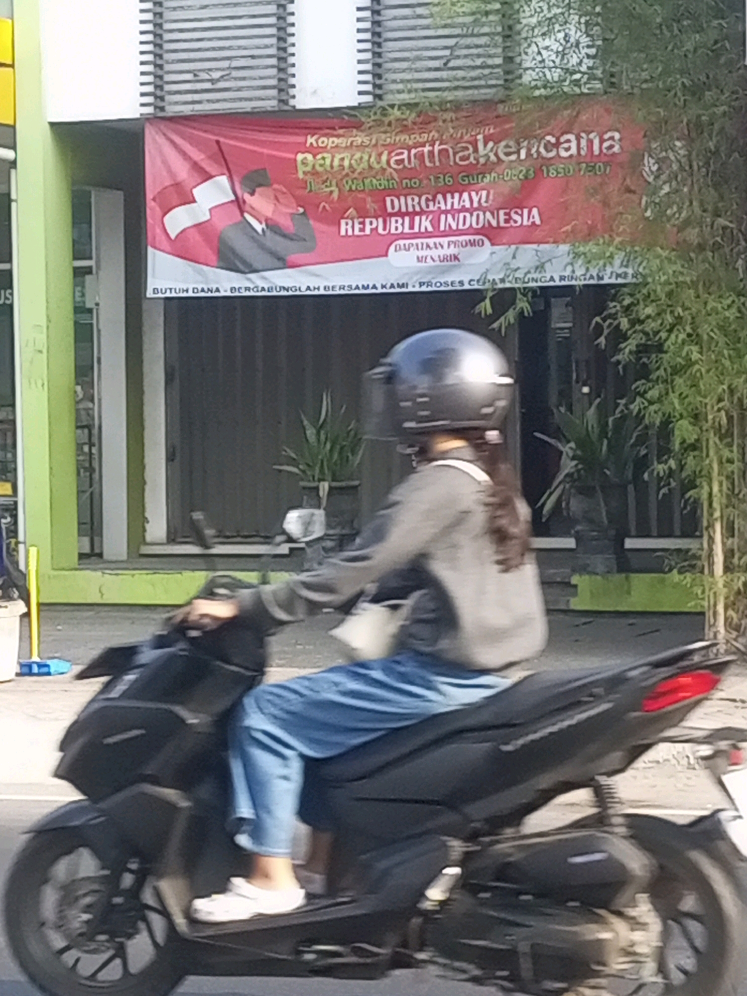 The image captures a person riding a black scooter on a street in Gurah, Indonesia. The rider, wearing a grey long-sleeved shirt, blue jeans, and a silver helmet, has long dark hair. They are also wearing white slip-on shoes. In the background, a red banner is prominently displayed. The banner features an illustration of a man in a suit saluting with the Indonesian flag. Text on the banner is in Indonesian and includes the words "Koprasi Simpan Pinjam Pangurthakencana," "DIRGAHAYU REPUBLIK INDONESIA," and the location "Gurah." The environment appears to be an urban or semi-urban setting with a building and greenery visible. The scooter's front wheel, handlebars, and the rider's legs are visible in the foreground.