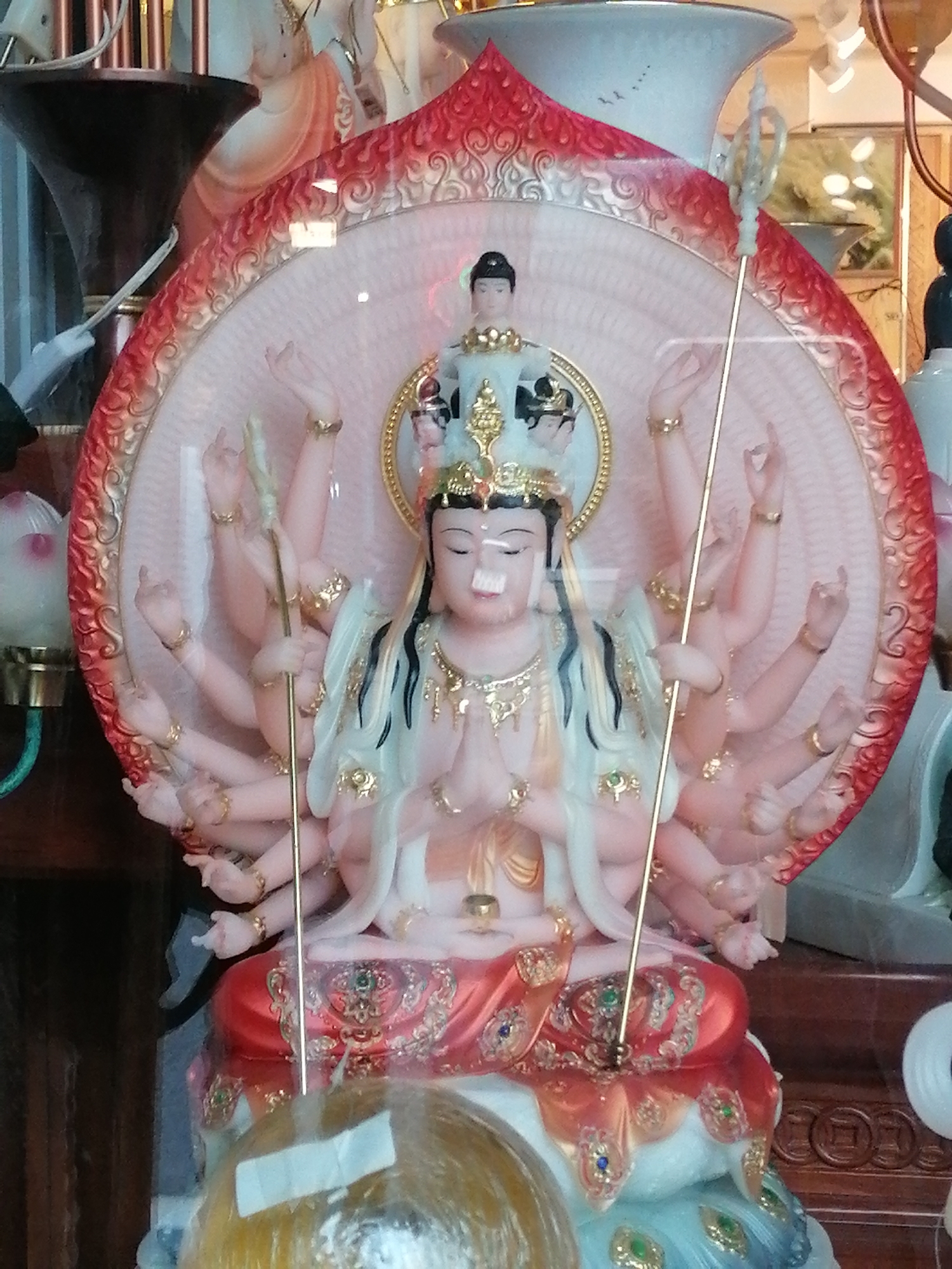 The image shows a statue of a multi-armed deity, likely Guan Yin, in a decorative setting. The statue is seated on a lotus-like base with intricate details and is surrounded by a fan-shaped backdrop adorned with red and gold accents. The deity has multiple arms, each with delicate hands. Some hands are in prayer or blessing gestures, while others hold objects. The deity wears a crown and ornate jewelry. The overall impression is of a religious or cultural artifact displayed indoors, possibly in a shop or temple in Da Lat, Vietnam. The background suggests an indoor environment with other objects and structures visible.