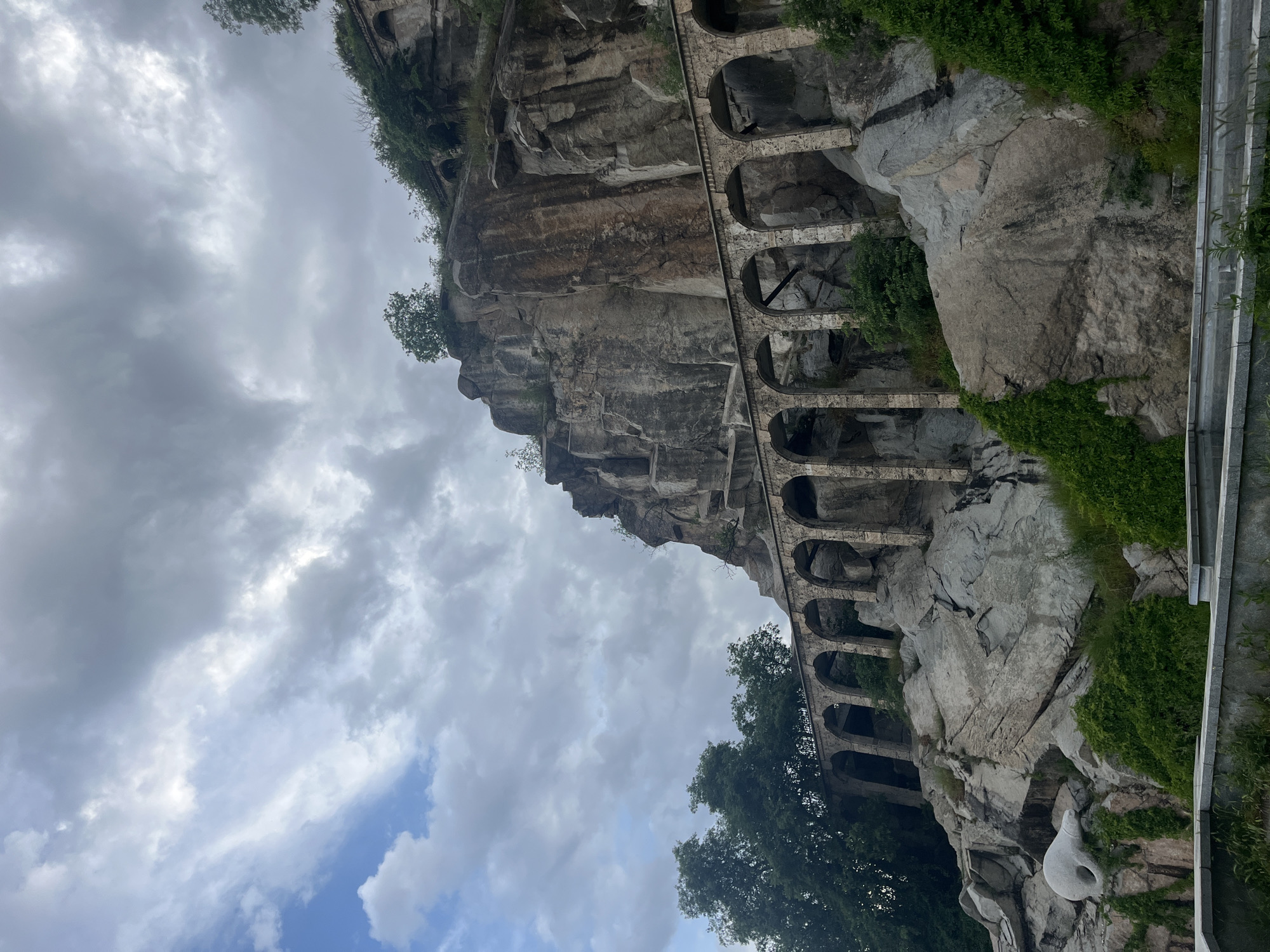 The image depicts an architectural structure built into the side of a cliff in Plovdiv, Bulgaria. This structure consists of a series of stone arches, creating a gallery or passage along the rock face. The cliff is primarily composed of grey rock and is partially covered with green vegetation. The sky is overcast with varying shades of grey clouds. In the foreground, there's some railing and what appears to be a modern art sculpture.
