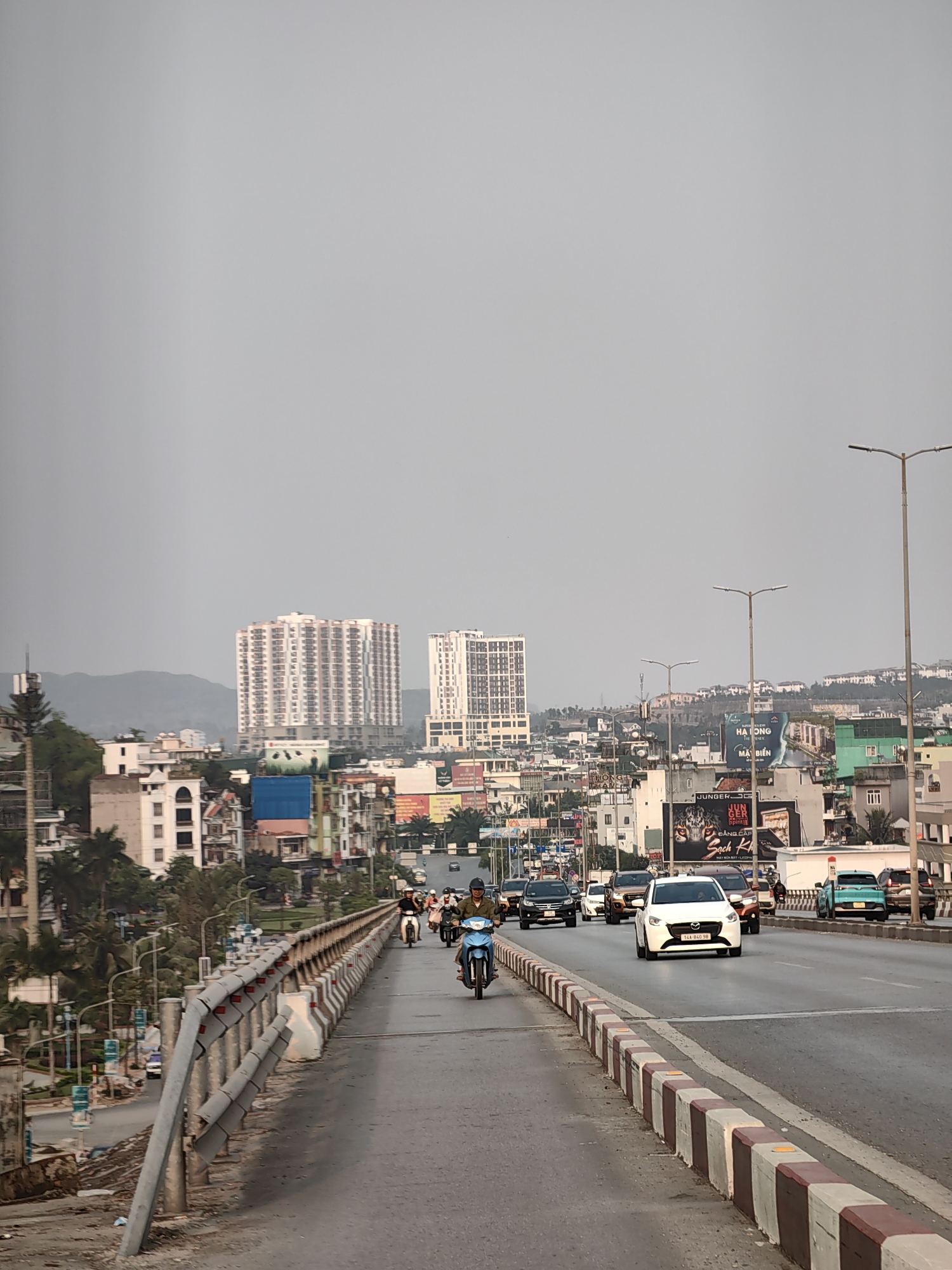 The image depicts a street scene in Hong Gai Ward, Vietnam, likely taken from an elevated position on a bridge or overpass. In the foreground, a multi-lane road stretches into the distance. Several vehicles are visible, including cars and motorbikes. People are seen riding the motorbikes and driving the cars. A white Mazda is the most prominent car in the image. Guardrails line the sides of the road with a patterned border of alternating white and brown. In the background, buildings and skyscrapers of varying heights are visible, along with billboards and signs in Vietnamese. The sky is overcast and grey. A mountainous terrain can be seen in the far distance. Streetlights stand tall on the right side of the road.