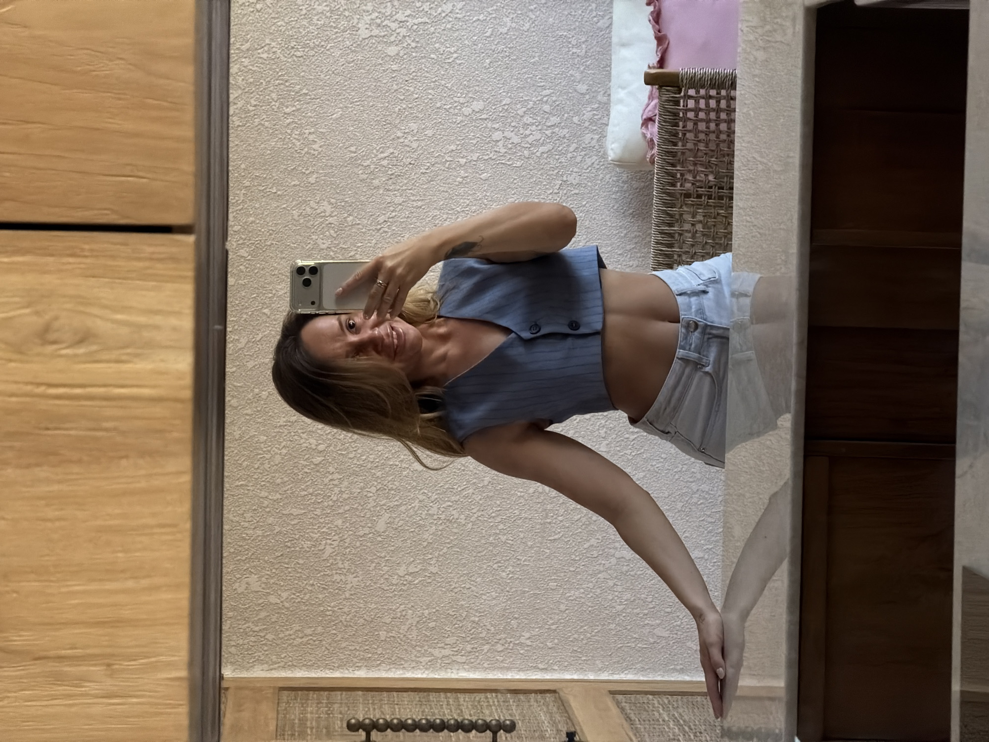 In the city of Mengwi, Indonesia, a woman with blonde hair takes a selfie in a mirror, wearing a light blue pinstriped vest and light-wash denim shorts. She is standing in what appears to be a hotel room, with a textured beige wall behind her and a wooden cabinet to her left. The mirror reflects a vanity with a marble top and dark wood cabinetry below. To the right, a woven chair with a cream-colored cushion and a pink throw are visible. The lighting suggests it is daytime.
