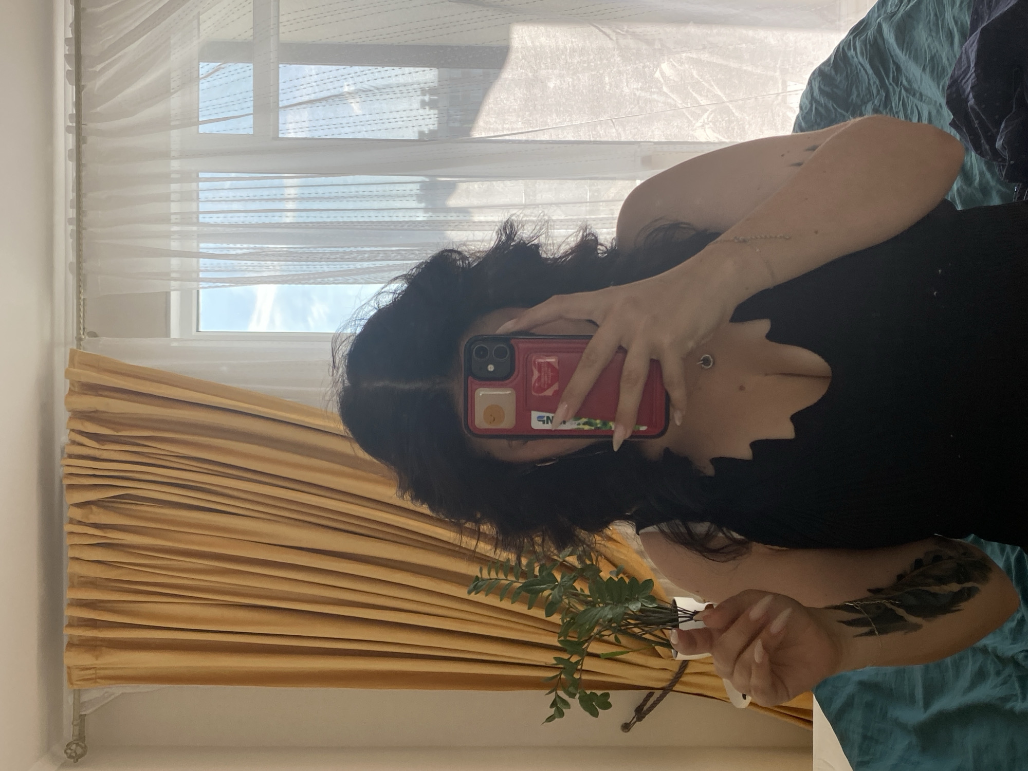 A young woman with dark, voluminous hair is taking a mirror selfie in a bedroom. She is wearing a black, low-cut tank top. Her right arm is raised, holding a red smartphone with a case that has stickers on it. A small potted plant with green leaves is visible in her left hand. She has a tattoo on her left arm, which appears to be black ink with feather-like designs. A delicate silver necklace with a small circular pendant is visible around her neck. She is standing in front of a window covered by sheer white curtains and thicker, golden-yellow drapes. The room is softly lit by natural light coming through the window. The background includes a bed covered with a teal-colored comforter. The overall setting suggests a casual, personal space. The image was taken in Yanino-1, Russia.