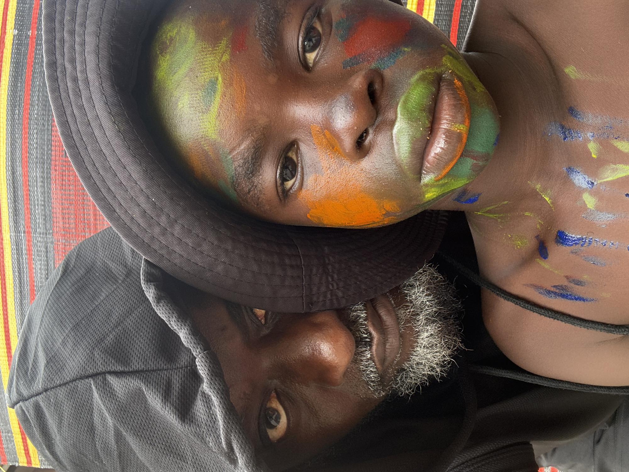 The image is a close-up, head-and-shoulders shot of two Black individuals, seemingly posing for a selfie. The person in the foreground is a child wearing a black bucket hat, their face and chest adorned with colorful paint. The paint is applied in broad strokes of green, orange, red, and yellow across the face, and blue and yellow on the chest and shoulder. The child's lips are prominently painted green and orange.
Partially obscured behind the child, on the left side of the frame, is an adult wearing a black hooded sweatshirt, with the hood pulled up. The adult's face is visible, with a grey beard and mustache. Their expression appears intense, with direct eye contact with the camera.
The background consists of a striped mat or fabric, with bands of grey, red, and yellow. The overall lighting suggests an outdoor setting. The image does not contain any elements that would indicate a specific location within the city of Ado, Nigeria.