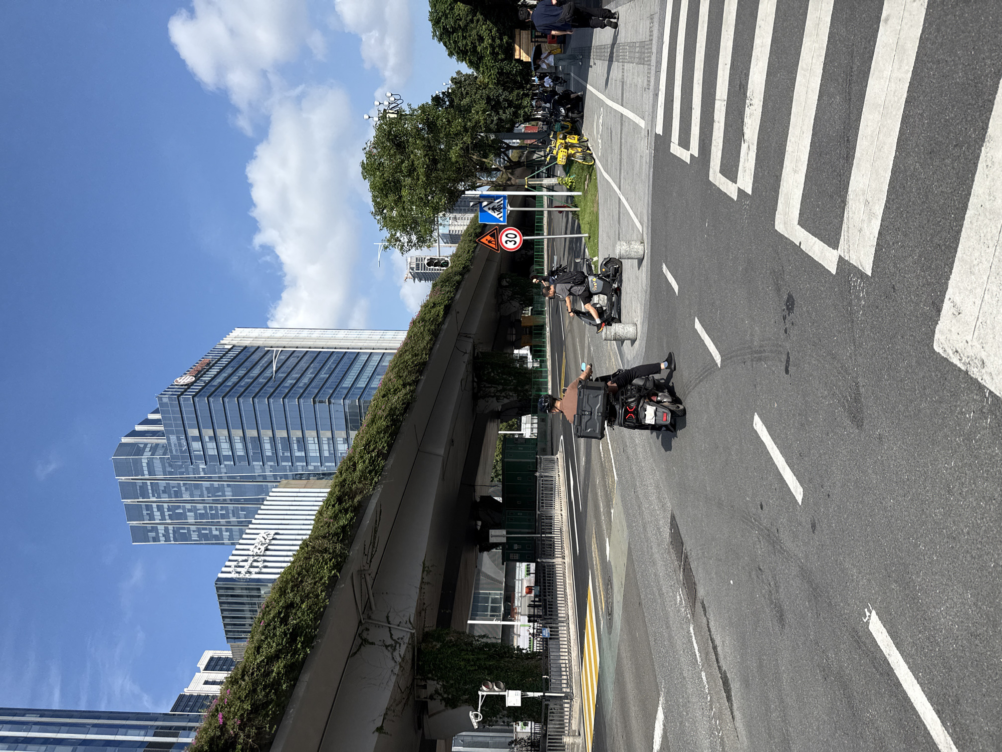 The image depicts a street scene in Tianhe District, China, with a large overpass covered in greenery dominating the upper left portion of the frame. Several modern, glass-fronted skyscrapers rise in the background. In the foreground, a two-lane road features a pedestrian crossing and lane markings. A person on a dark-colored electric scooter, equipped with a large black storage box on the rear, is riding in the right lane. Another person on a similar scooter is positioned further ahead in the same lane. A yellow bicycle is parked on the sidewalk to the right of the road, alongside other parked vehicles. Traffic signs, including a speed limit sign of "30" and pedestrian crossing symbols, are visible on the right side of the road. The sky is blue with scattered white clouds.