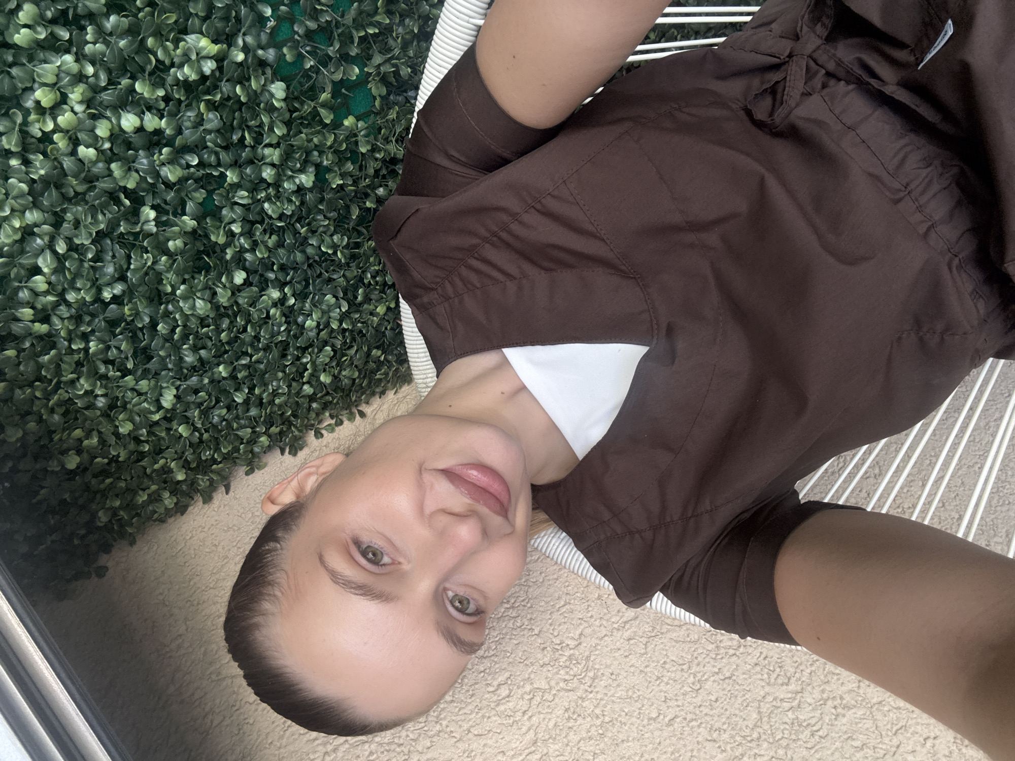 A woman with her hair pulled back in a ponytail, wearing a brown scrub top over a white shirt, sits on a white chair. She is looking at the camera and smiling slightly. Behind her is a wall of green artificial foliage, and to her left is a textured beige wall. The scene appears to be outdoors, possibly on a balcony or patio, with a metallic overhang visible at the top left. The context of Budva, Montenegro is not discernable from the visual information in the image.