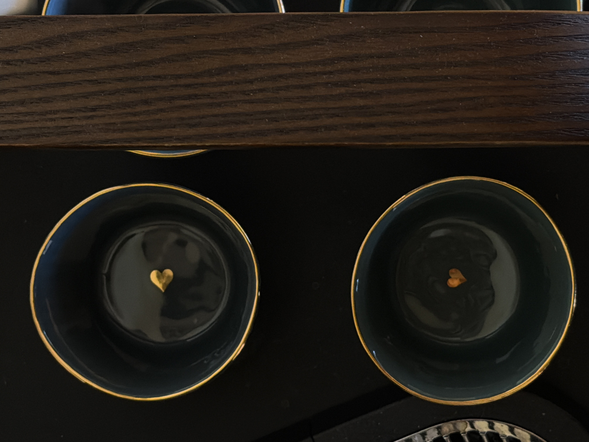 The image shows two dark teal teacups with gold rims. Each cup has a small gold heart at the bottom inside. The cups are placed on a black surface, with a dark brown, likely wooden, object above them. There's a glimpse of a silver, possibly metal, grate in the bottom right corner. The setting appears to be indoors, possibly a kitchen or dining area. No people are visible. The location in Riga, Latvia is not discernible from the image.