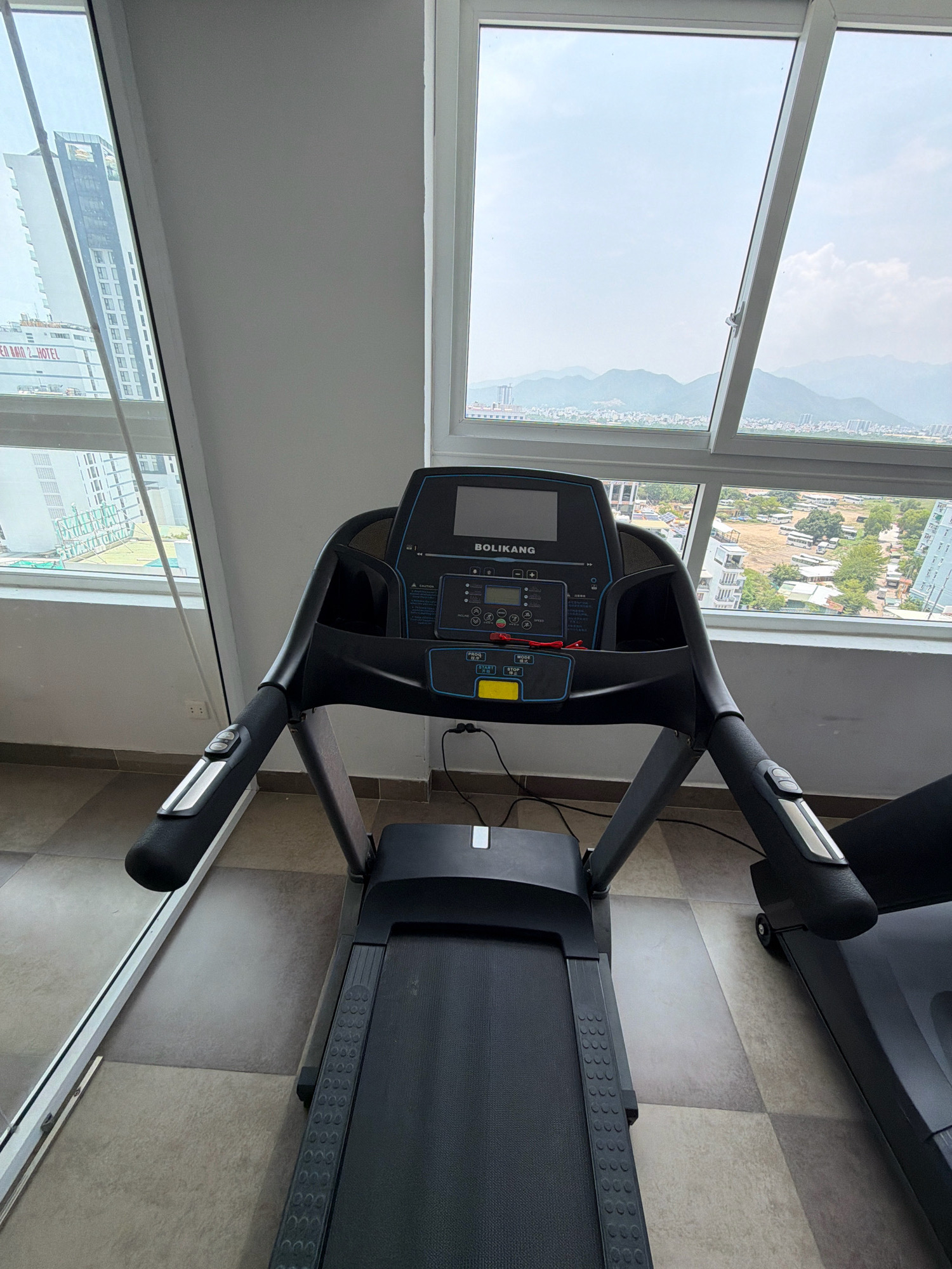 The image shows a treadmill in a room with large windows overlooking a city and mountains. The treadmill is black with a black running belt and silver accents on the handles. The console has a digital display and buttons. The room has tiled flooring. Outside the window, there are buildings, trees, and a hazy sky. The location is identified as Nha Trang, Vietnam, with a sign visible on a distant building that appears to say "BIEN NAM 2 HOTEL". There are no people or actions depicted in the image.