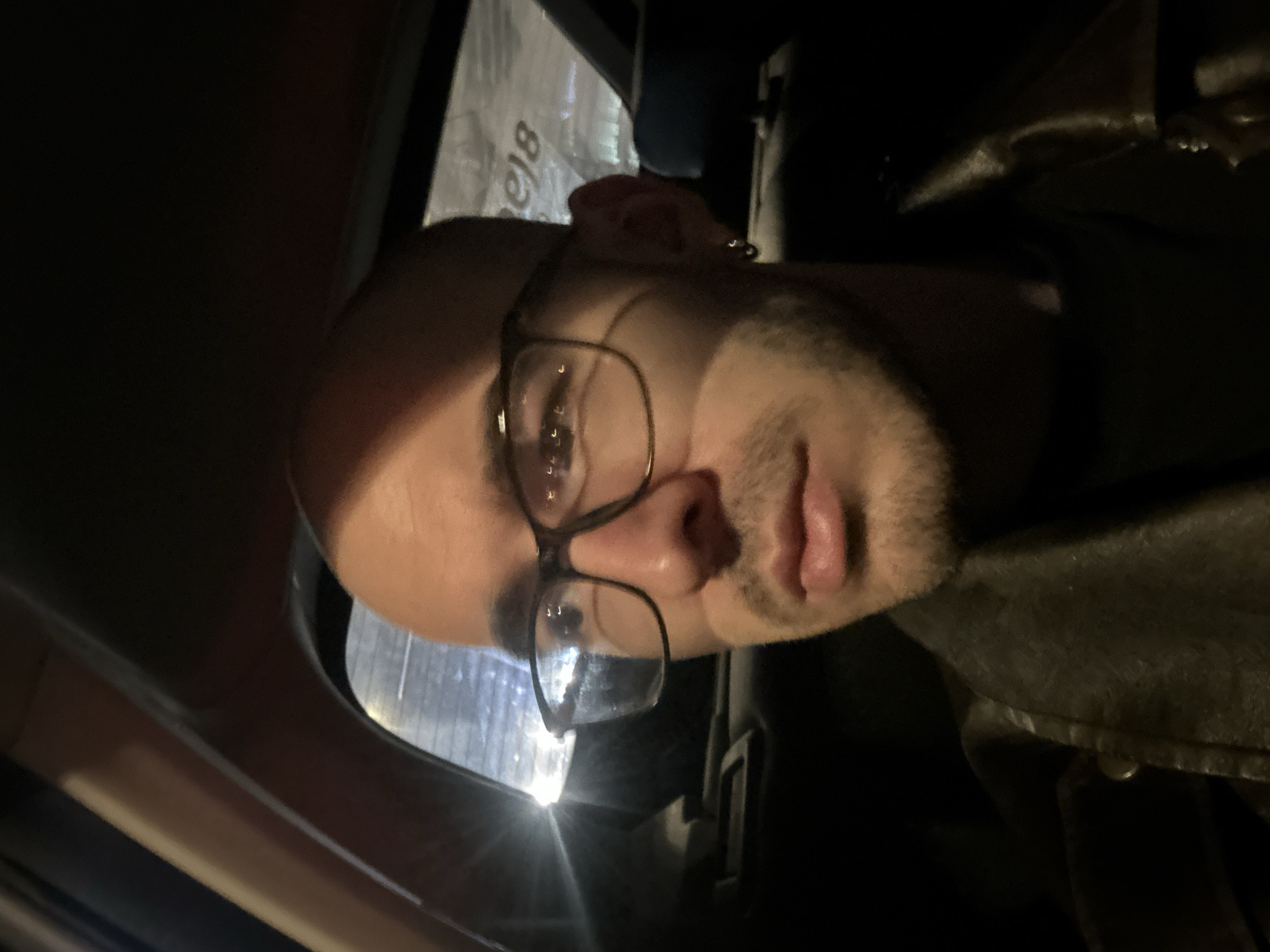 A close-up, low-light photograph shows a person wearing glasses with a shaved head inside a vehicle, possibly a car. The person is positioned at an angle, looking into the camera. The interior of the vehicle is dark, with a glimpse of what appears to be a window or a reflective surface in the background, exhibiting some light flare. The subject is wearing a dark jacket. The location is indeterminate; there is insufficient detail to determine if the photo was taken in Moscow, Russia.