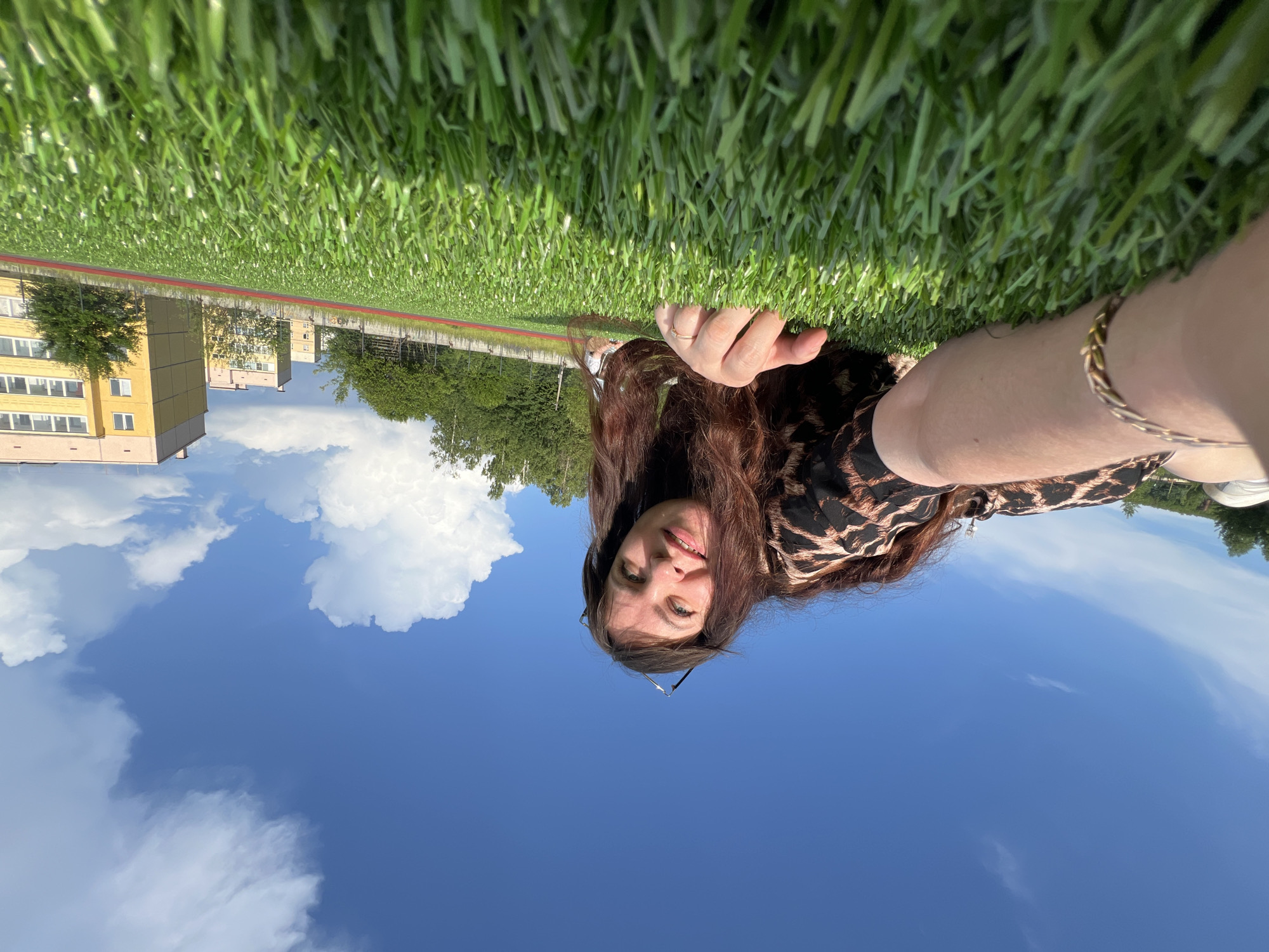 A woman with long, brown hair is lying on a vibrant green artificial turf field, looking up at the blue sky with white clouds. She is wearing a leopard-print dress and a bracelet on her left wrist. Her right hand is resting on the grass. In the background, there are trees, a red running track, and several residential buildings with yellow and beige facades, consistent with the architecture found in Noviy, Russia. The low-angle shot emphasizes the expanse of the sky and the texture of the artificial grass.