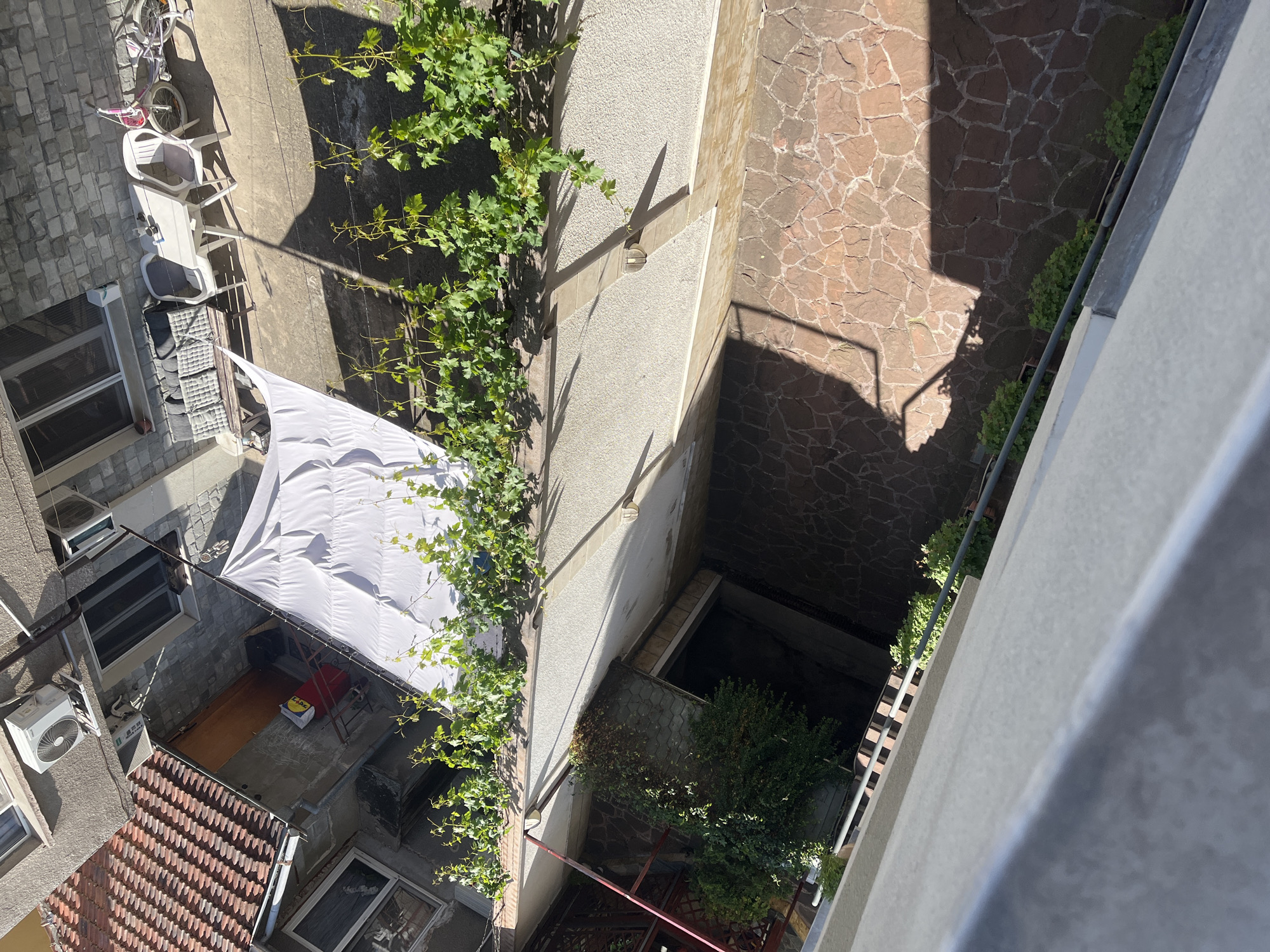 This overhead, high-angle photograph captures a narrow outdoor space, likely a courtyard or alleyway, situated between urban buildings. The scene is bathed in bright sunlight, casting sharp, distinct shadows that suggest it is midday or early afternoon with clear skies.
On the left, a paved area featuring irregular grey stone tiles is visible at the top, where a pink and white bicycle is parked. Below this, a white plastic table and chairs are arranged. Further down the left building facade, several air conditioning units protrude from the wall above windows. A large white tarp or awning extends over a lower section of the courtyard, providing shade. Beneath this tarp, an outdoor area with a light-colored floor, a red metal frame, and a yellow bag (with indistinct text, possibly "LUX") can be seen. Below this is a reddish-brown tiled roof.
The central part of the image is dominated by a light-colored building wall, extensively covered by climbing green vine plants. This wall casts prominent shadows onto the ground and adjacent structures. The ground level transitions from paved sections to a deeper, darker area with some small green bushes.
To the right, a plain grey building wall stands beside a ground area paved with dark, irregular stone tiles. Shadows stretch across this tiled surface. A thin line of green vegetation runs along the base of the wall on the right. No people or specific activities are visible. The setting is characteristic of an urban environment, consistent with the provided location context of Sofia, Bulgaria.