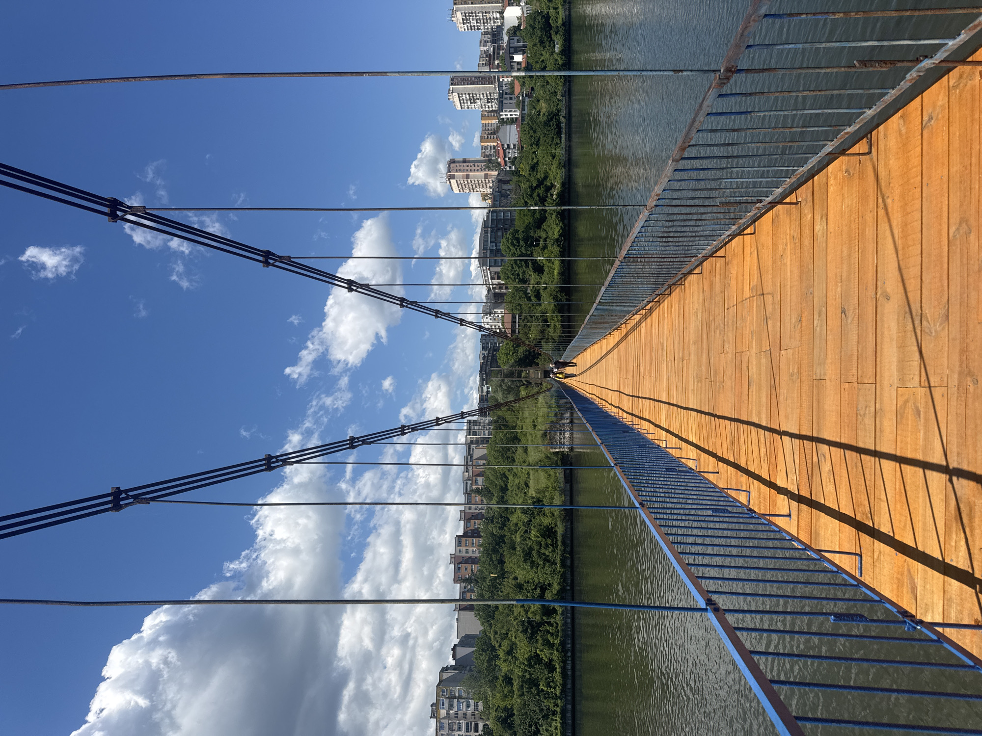 Here is a detailed description of the image:
The image shows a straight shot down the middle of a pedestrian suspension bridge. The bridge has a wooden plank surface stained with a warm orange color. On either side of the bridge, blue-painted metal railings run the length. Overhead, suspension cables are visible against a bright blue sky dotted with fluffy white clouds. The cables attach to the bridge structure at regular intervals.
Below the bridge, a river flows, with green, tree-lined banks on both sides. Buildings and city infrastructure are visible further back from the riverbanks.
The perspective of the photograph makes the bridge appear to stretch into the distance, converging towards the center of the frame. The image is rotated 90 degrees clockwise.