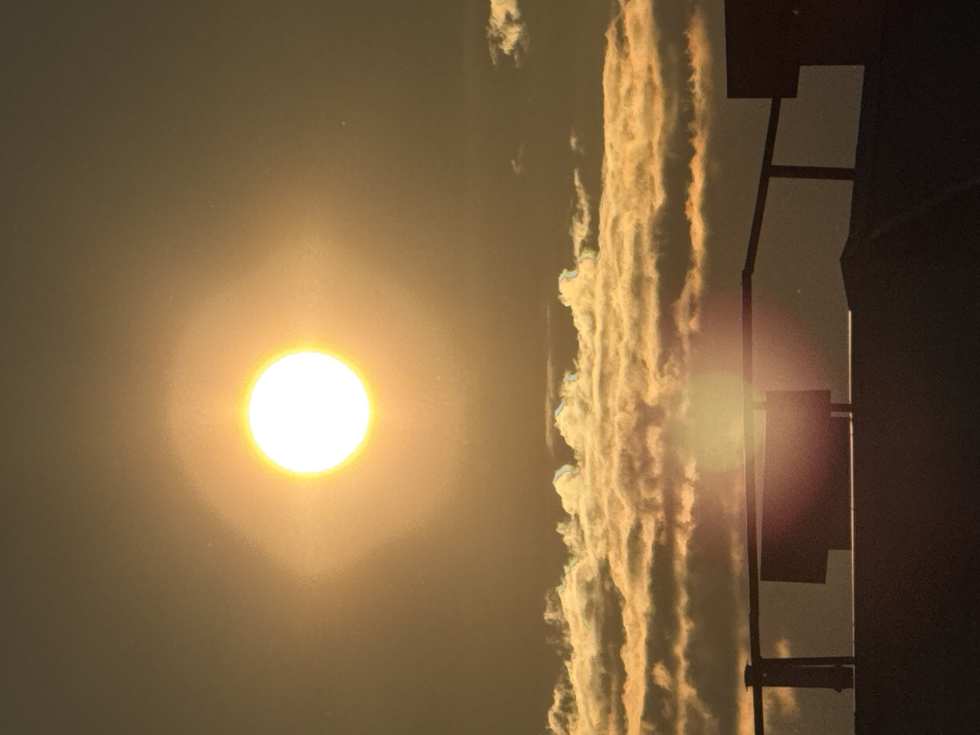 The image displays an atmospheric sky scene, dominated by a bright, circular sun positioned prominently on the left side. The sun radiates an intense white and yellow glow, illuminating the surrounding hazy sky with warm golden and ochre tones.
Stretching horizontally across the middle and right portions of the frame are distinct formations of clouds. These clouds appear as elongated, textured bands, brightly illuminated by the sun's rays. Their surfaces showcase vibrant shades of golden yellow, white, and light orange, creating a striking contrast against the darker parts of the sky.
On the far right, dark, sharp silhouettes of structures are visible. These structures feature angular lines and solid shapes, suggesting elements of a building, such as a window frame, a railing, or a light pole, set against the illuminated sky. A subtle lens flare is also apparent on the right, overlaying some of the silhouetted structures.
The scene depicts an outdoor setting, likely at either sunrise or sunset, given the warm hues and the low perceived position of the sun relative to the horizon (if the image were rotated). The weather appears to be cloudy and somewhat hazy. No people are present in the image. The location context for this scene is Markovskiy, Russia. There is no visible text.